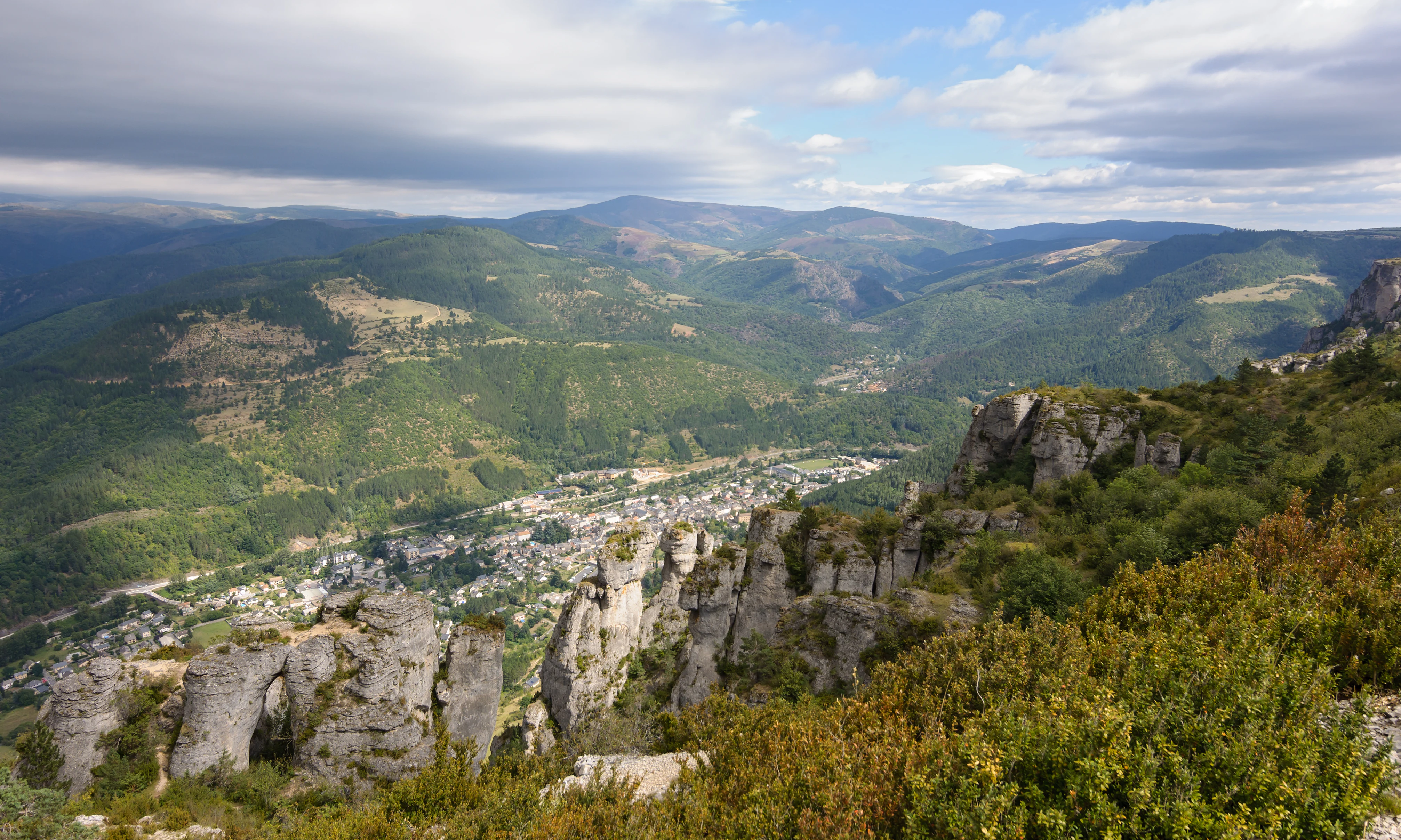 Wide landscape view of a mountain valley with rocky outcrops, a town below, and distant mountain ranges under a partly cloudy sky