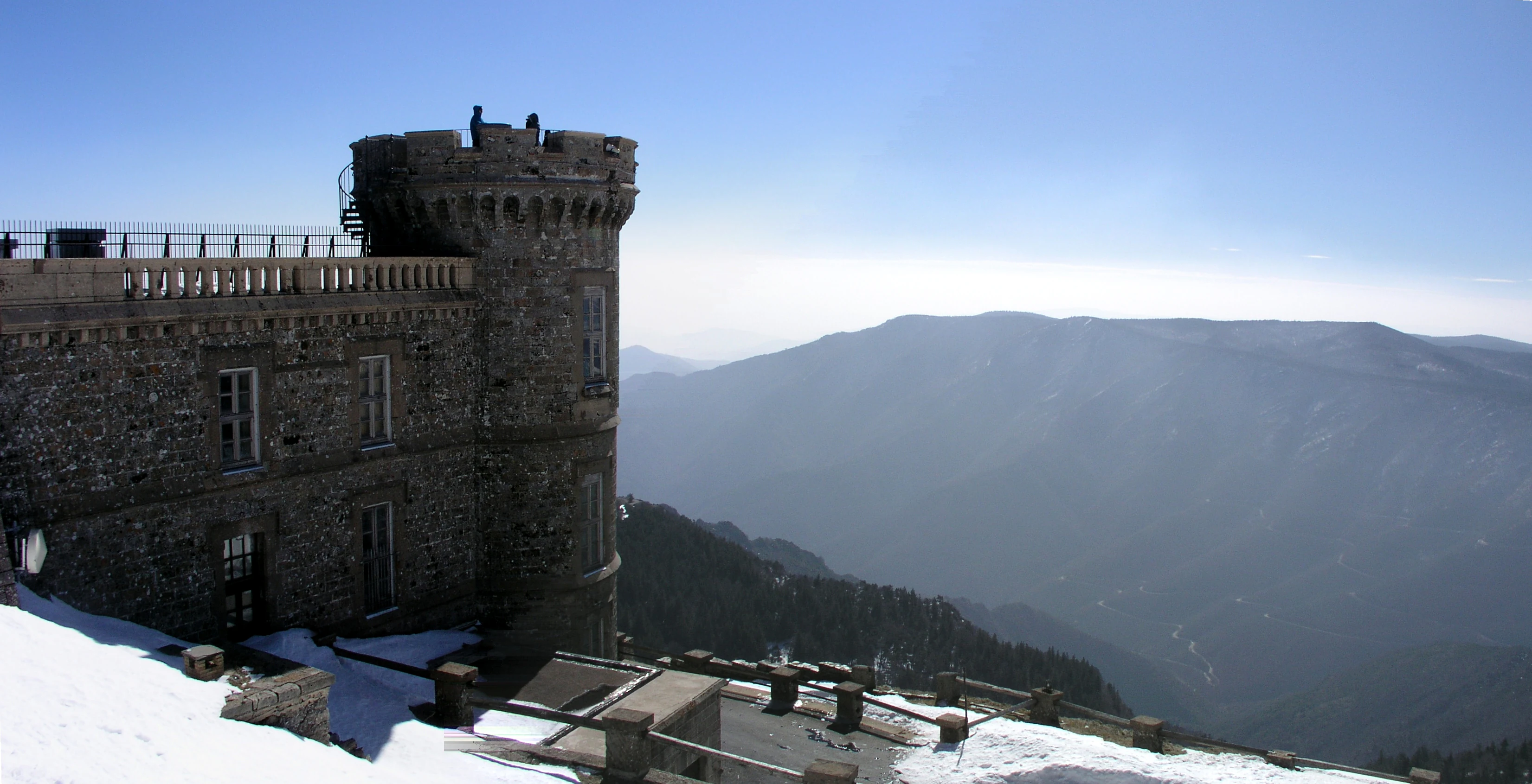 Stone tower building with snow on ground, mountains in background, clear blue sky