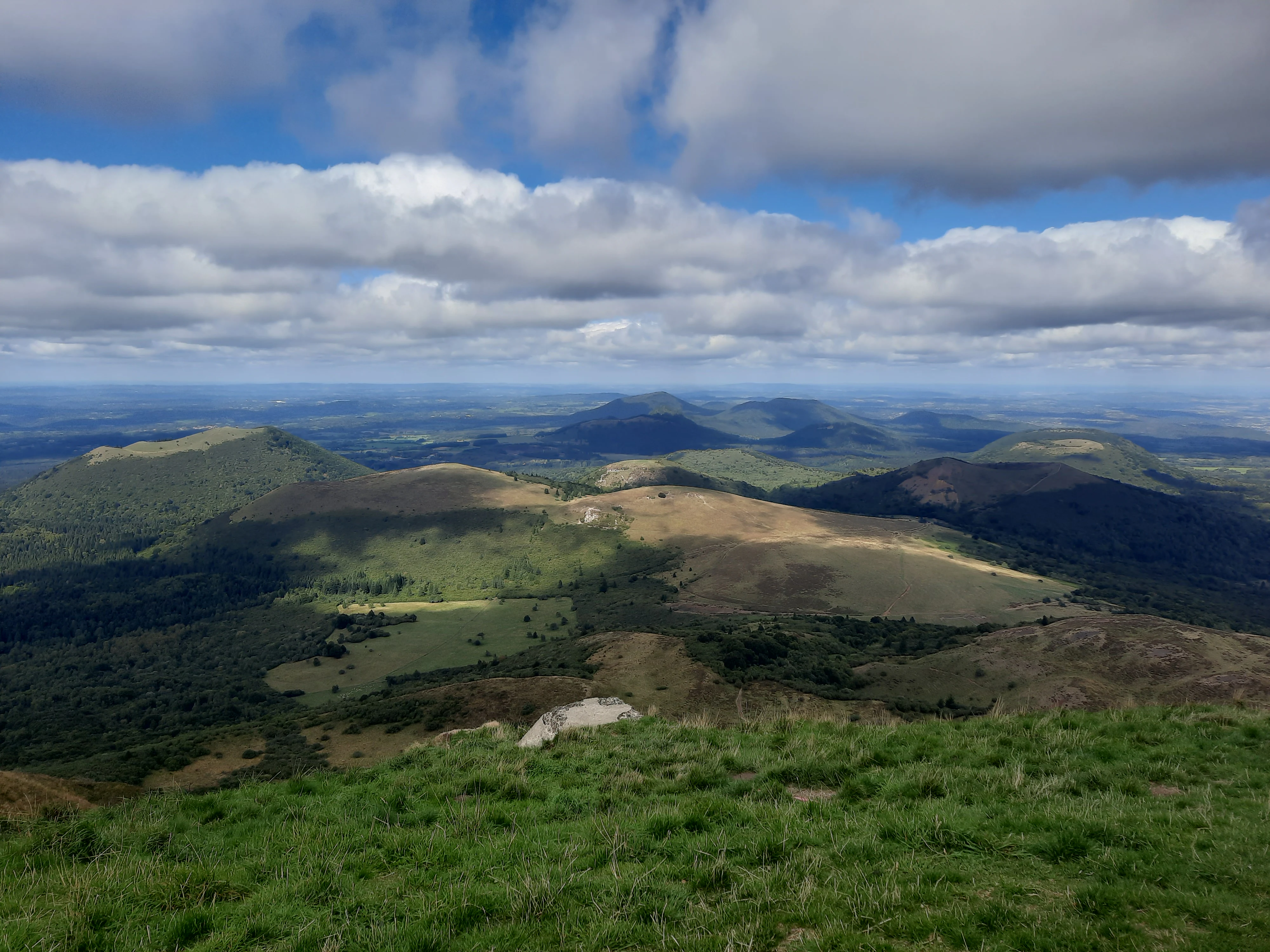 Wide-angle landscape view from a high mountain ridge looking over multiple volcanic peaks and rolling green hills under a cloudy sky.