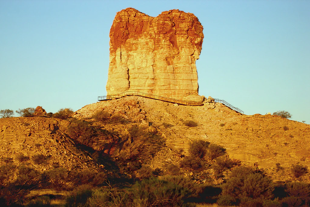 Un gran pilar de arenisca que se eleva sobre un paisaje desértico rocoso al atardecer, iluminado por una cálida luz anaranjada.