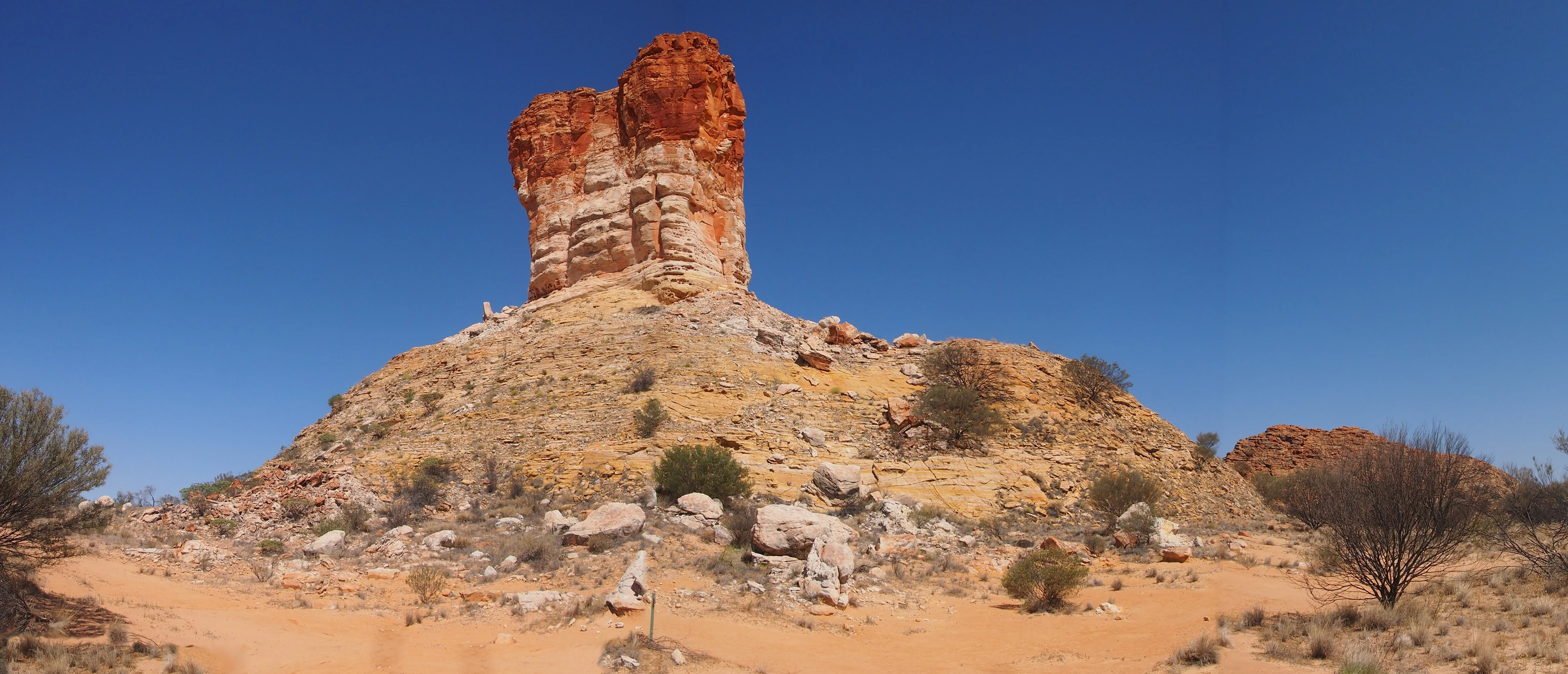 A wide angle view of the sandstone rock formation Chambers Pillar rising above a desert landscape under a clear blue sky.