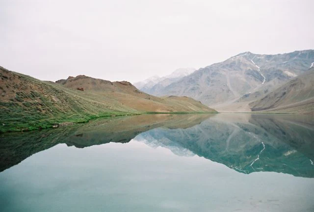 A wide, calm, turquoise high-altitude lake reflecting surrounding barren, rocky mountain slopes under an overcast sky.
