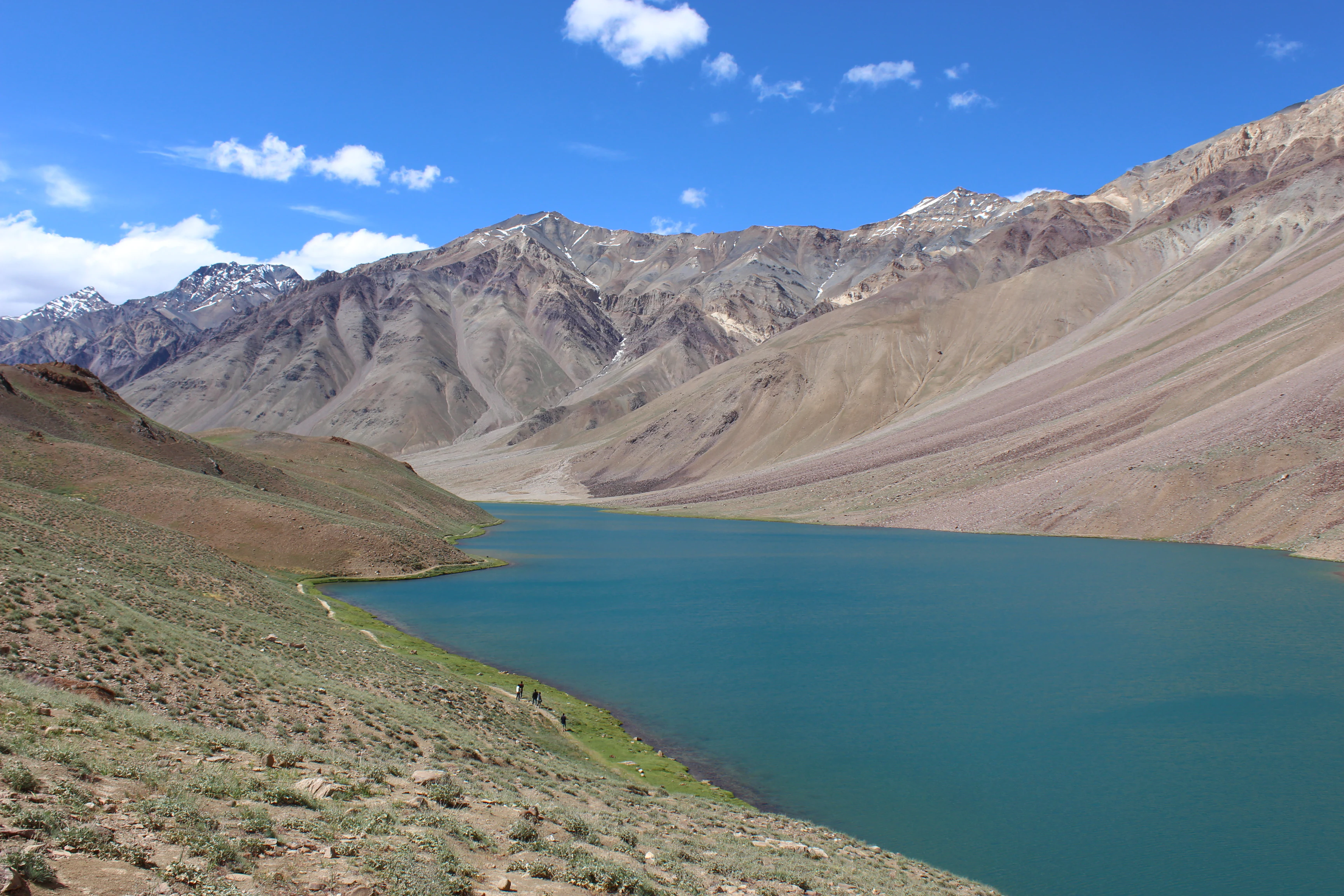 A wide landscape photograph of a turquoise lake nestled within a deep valley, surrounded by barren, brown Himalayan mountain slopes under a clear blue sky.