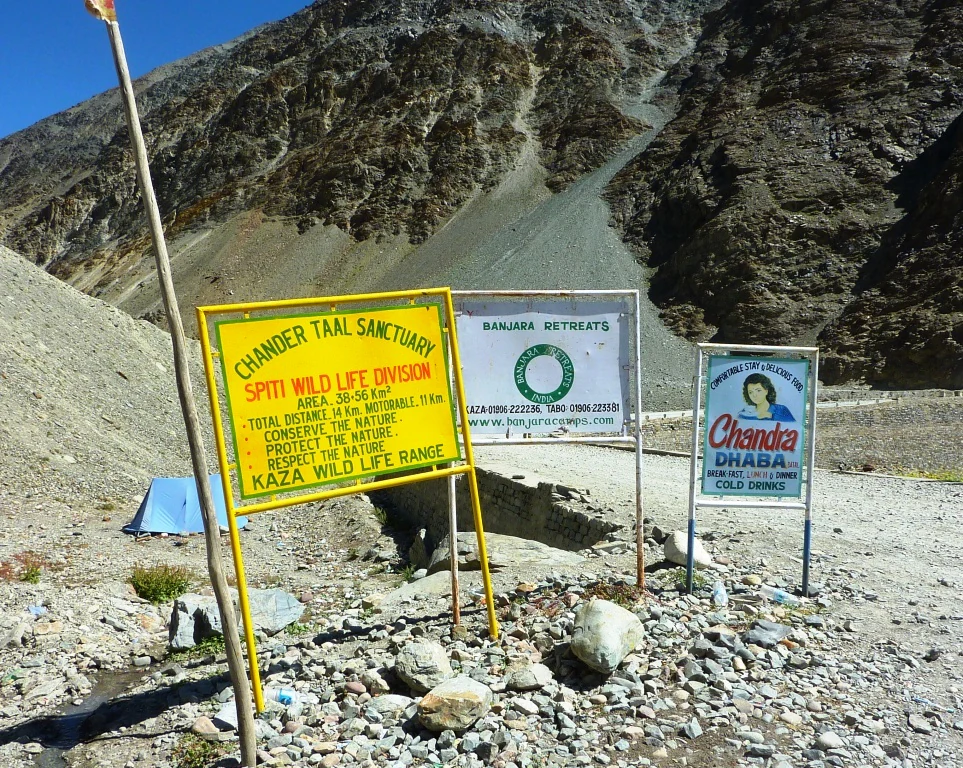 Three metal signs standing on a gravel ground with a rocky, arid mountain slope in the background. The leftmost sign is a yellow board labeled Chander Taal Sanctuary with conservation text.