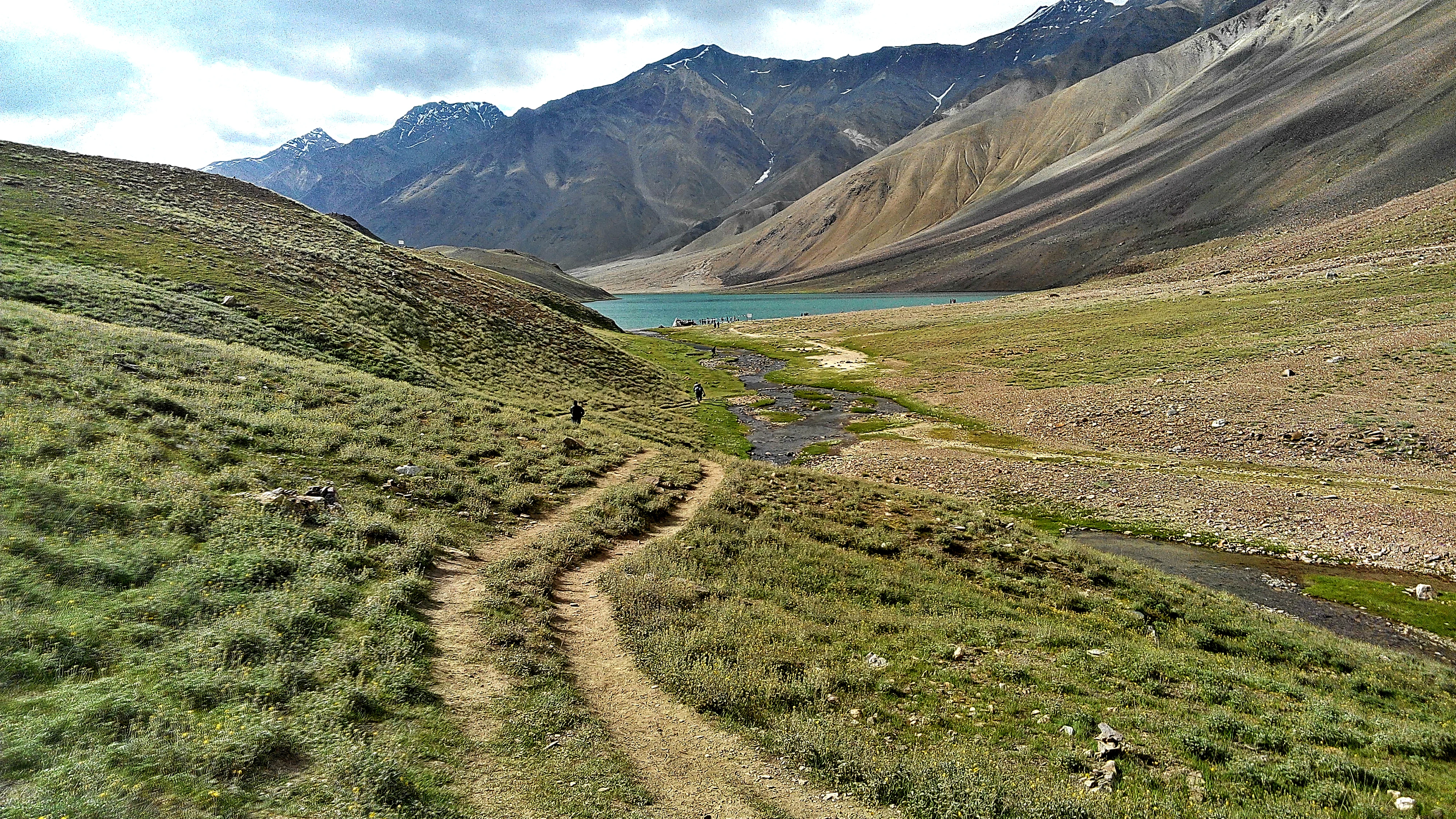 A dirt path leads through green alpine meadows toward a crescent-shaped mountain lake surrounded by rugged, rocky Himalayan peaks.