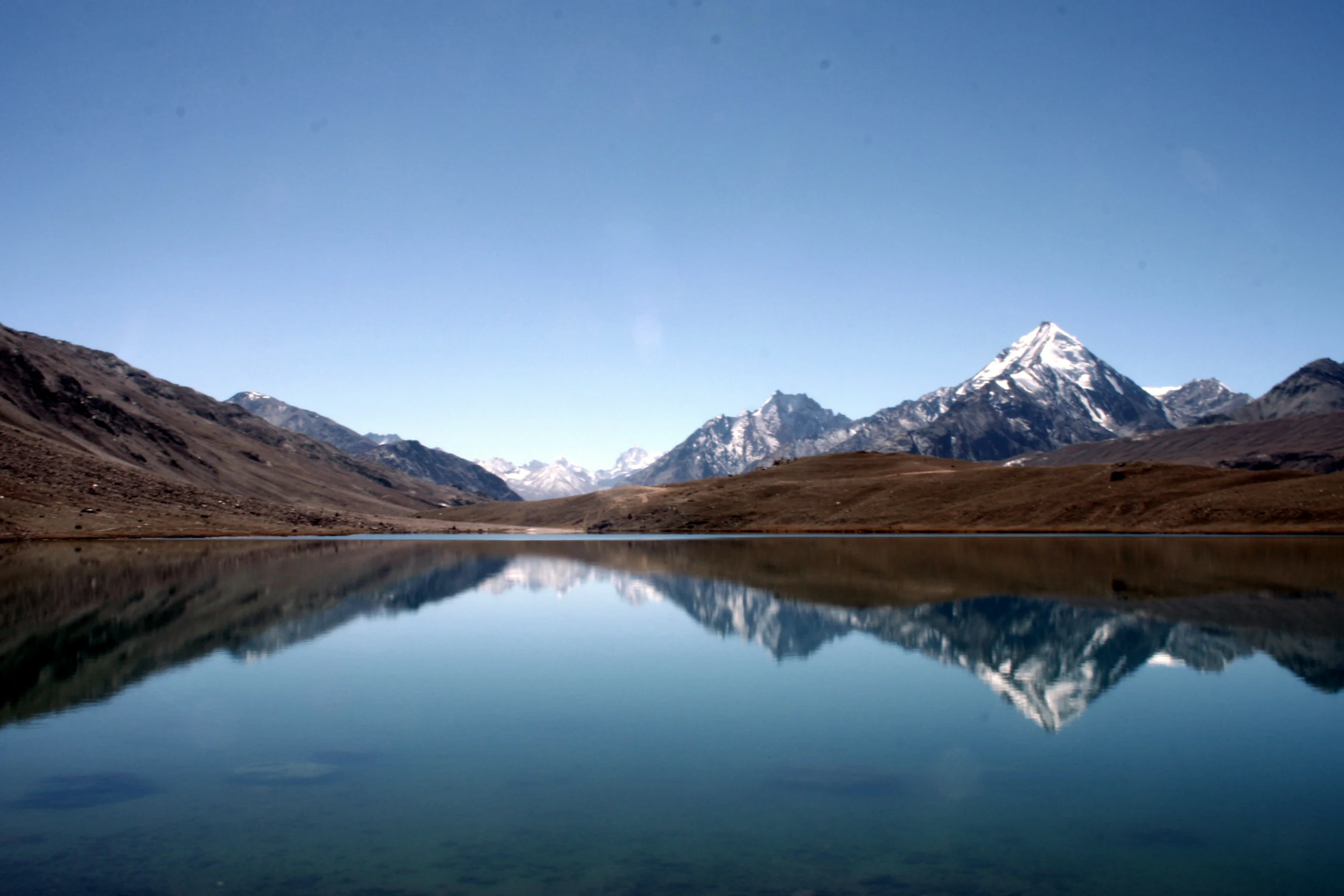 A clear, calm high-altitude lake reflects the surrounding barren mountains and a snow-capped peak under a bright blue sky.