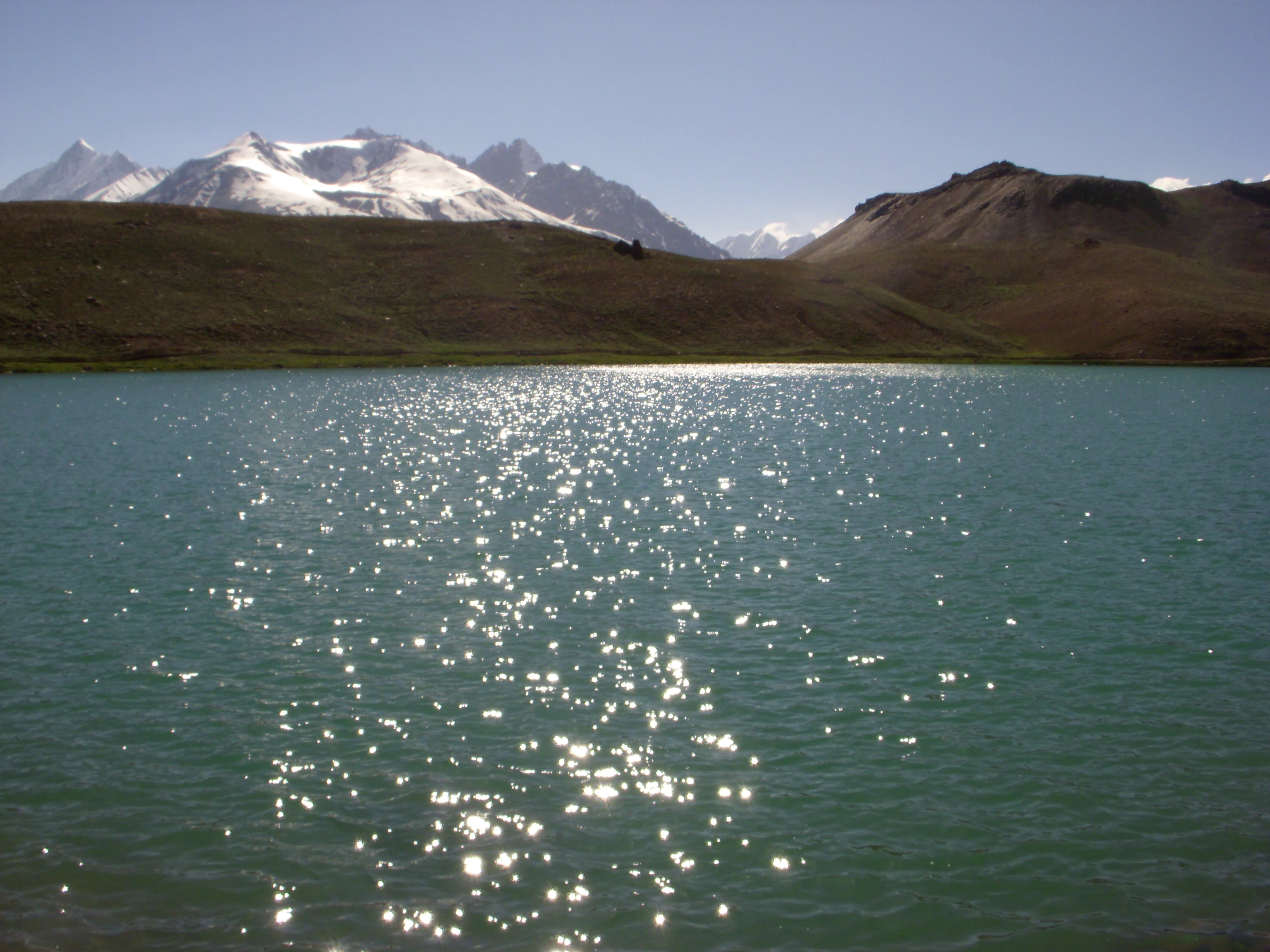 A bright, turquoise mountain lake with sunlight reflecting on the water, framed by snow-capped peaks and barren brown hills under a clear blue sky.