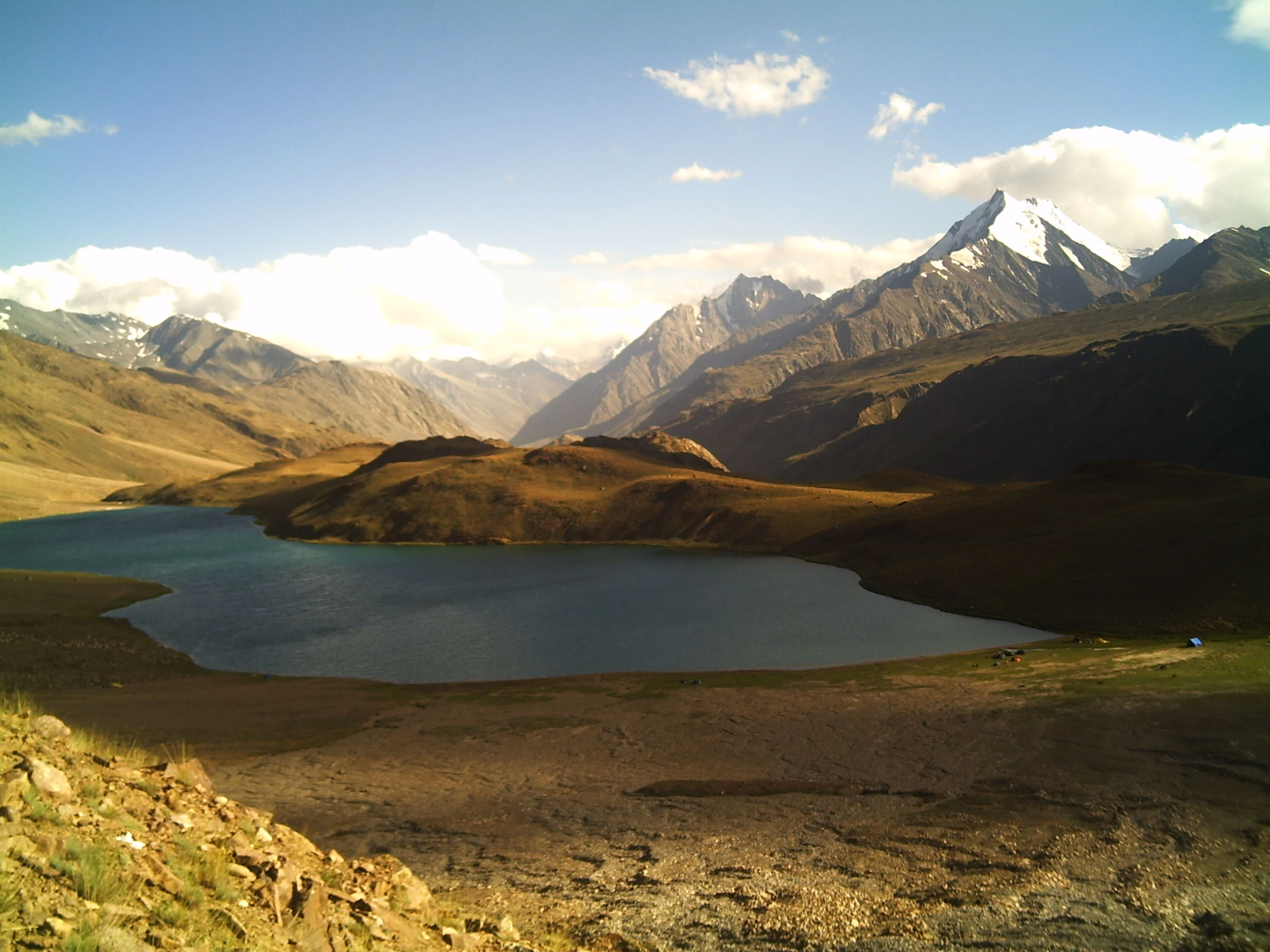 A wide angle view of a blue alpine lake set in a valley, with snow-capped mountain peaks rising in the background under a blue sky with white clouds.