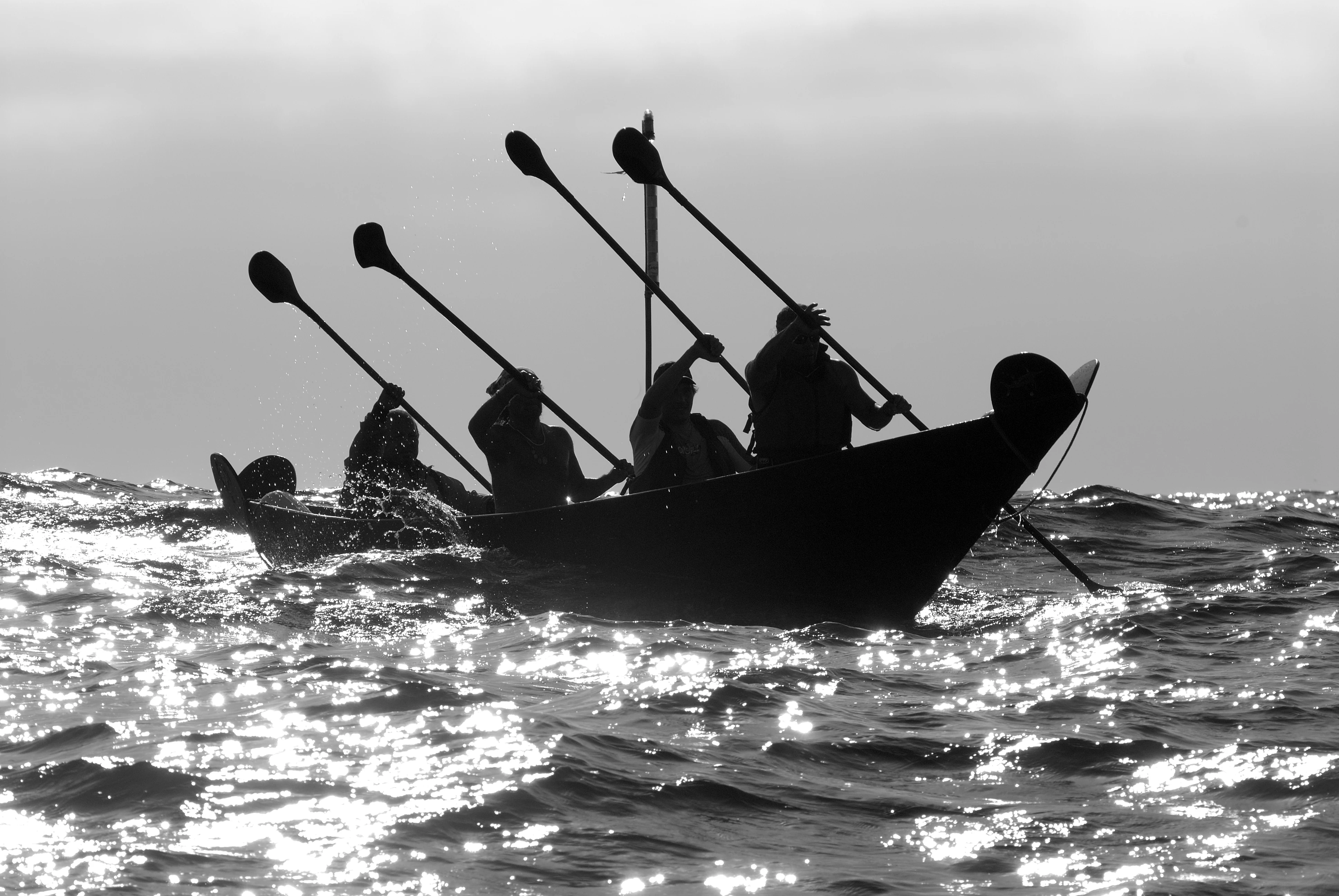 A black and white silhouette of a traditional Chumash plank canoe with four paddlers navigating choppy, sun-glinting ocean waters.
