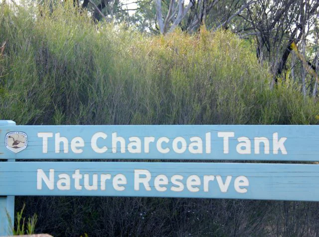 A light blue wooden sign reading The Charcoal Tank Nature Reserve in white text, situated in front of dense green scrub and trees.