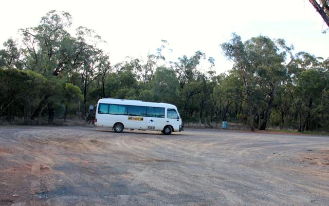 A small white bus parked in a large dirt clearing surrounded by native eucalyptus forest.