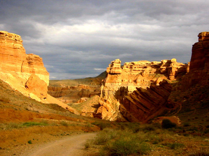 A dirt path leading into a deep canyon with towering red sandstone cliffs under a cloudy sky.