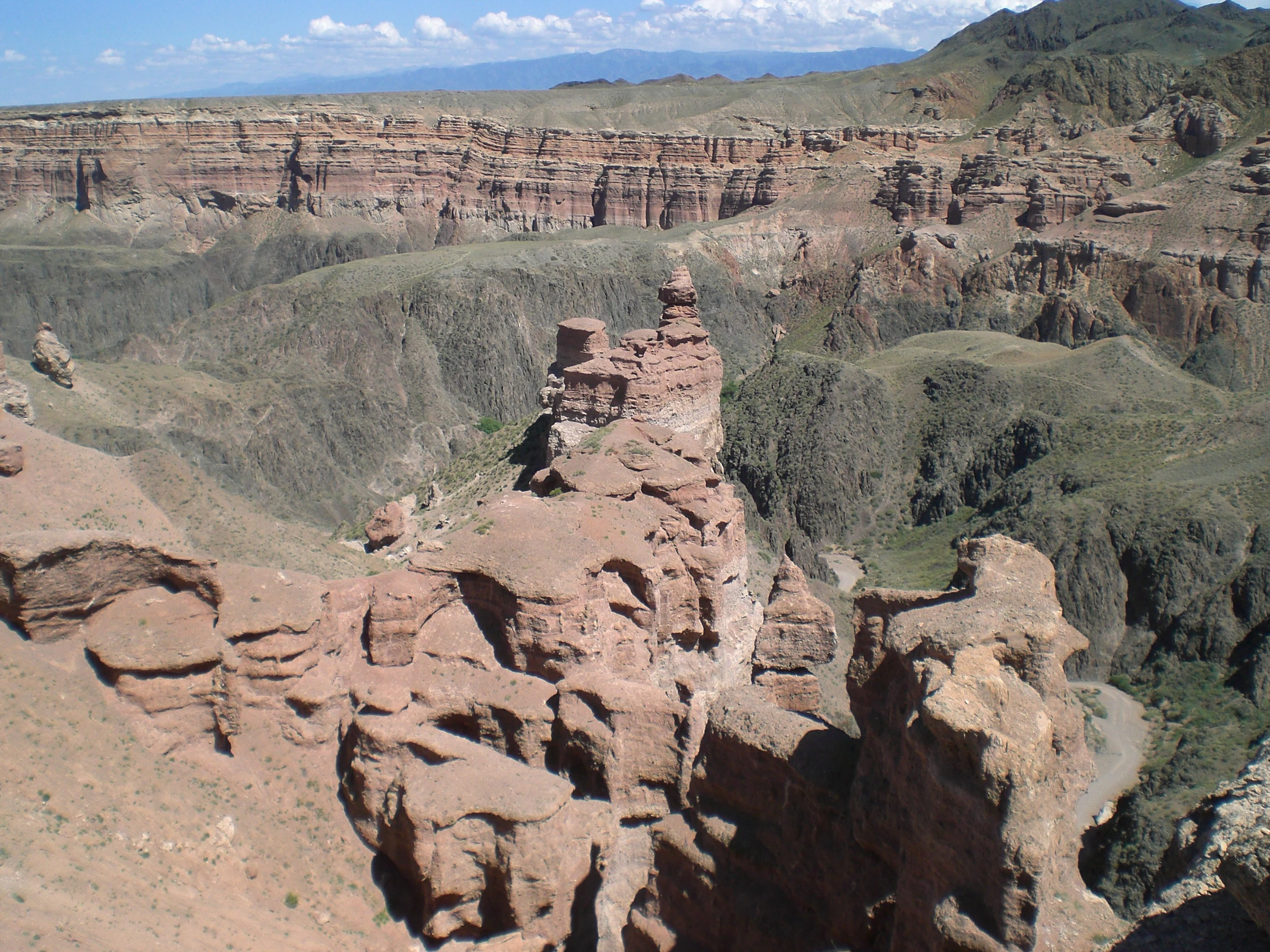 Red rock formations and canyon landscape with layered cliffs and green terrain under a partly cloudy sky
