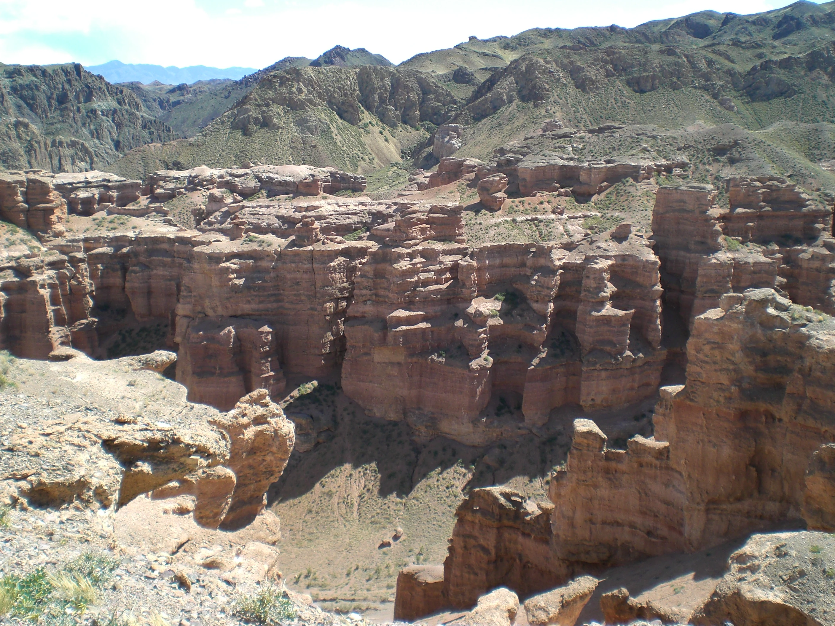 Red rock canyon landscape with steep cliffs, rocky formations, and distant mountains under a partly cloudy sky