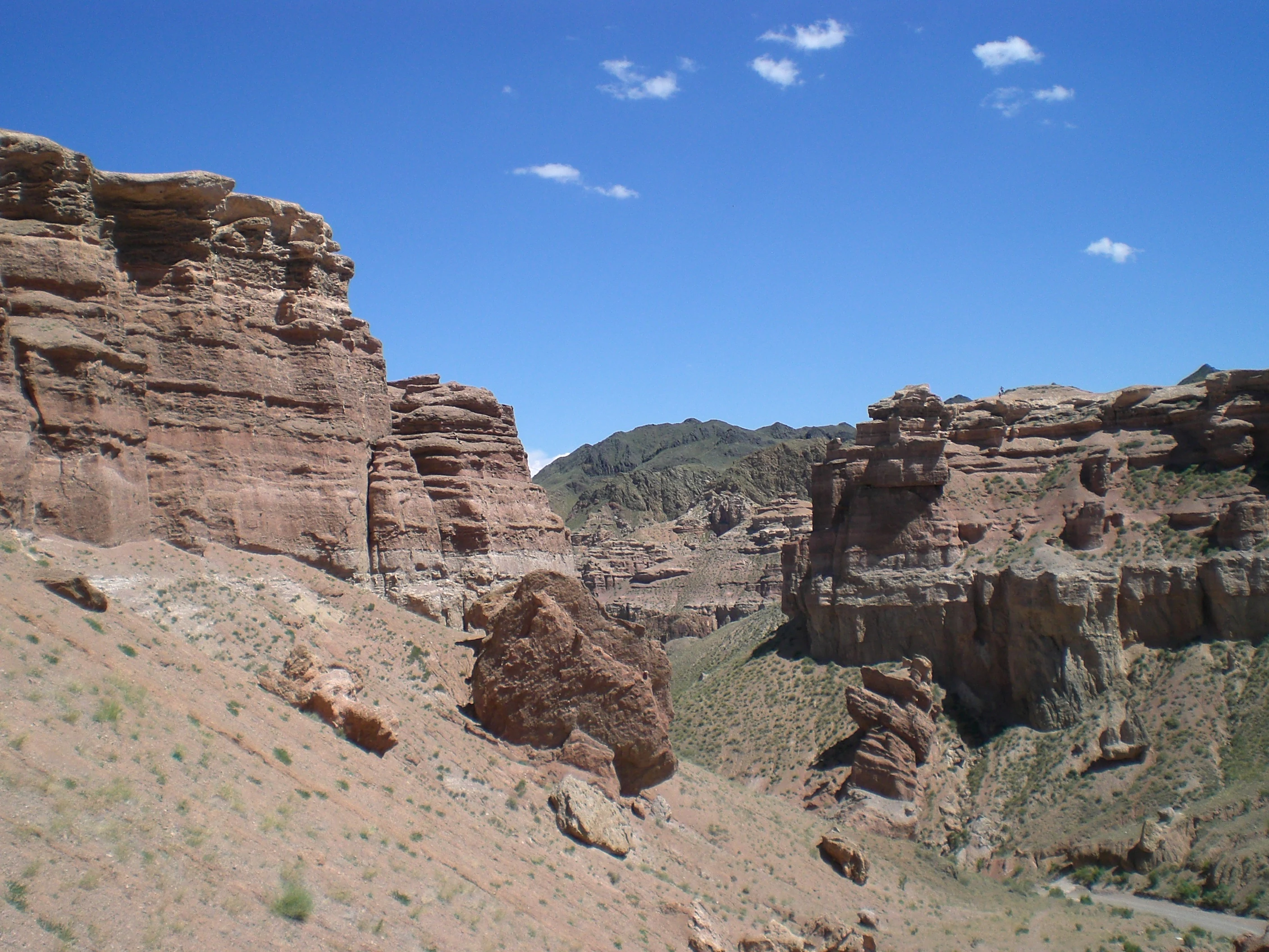 Rocky canyon slopes with layered red rock formations under a clear blue sky with scattered clouds