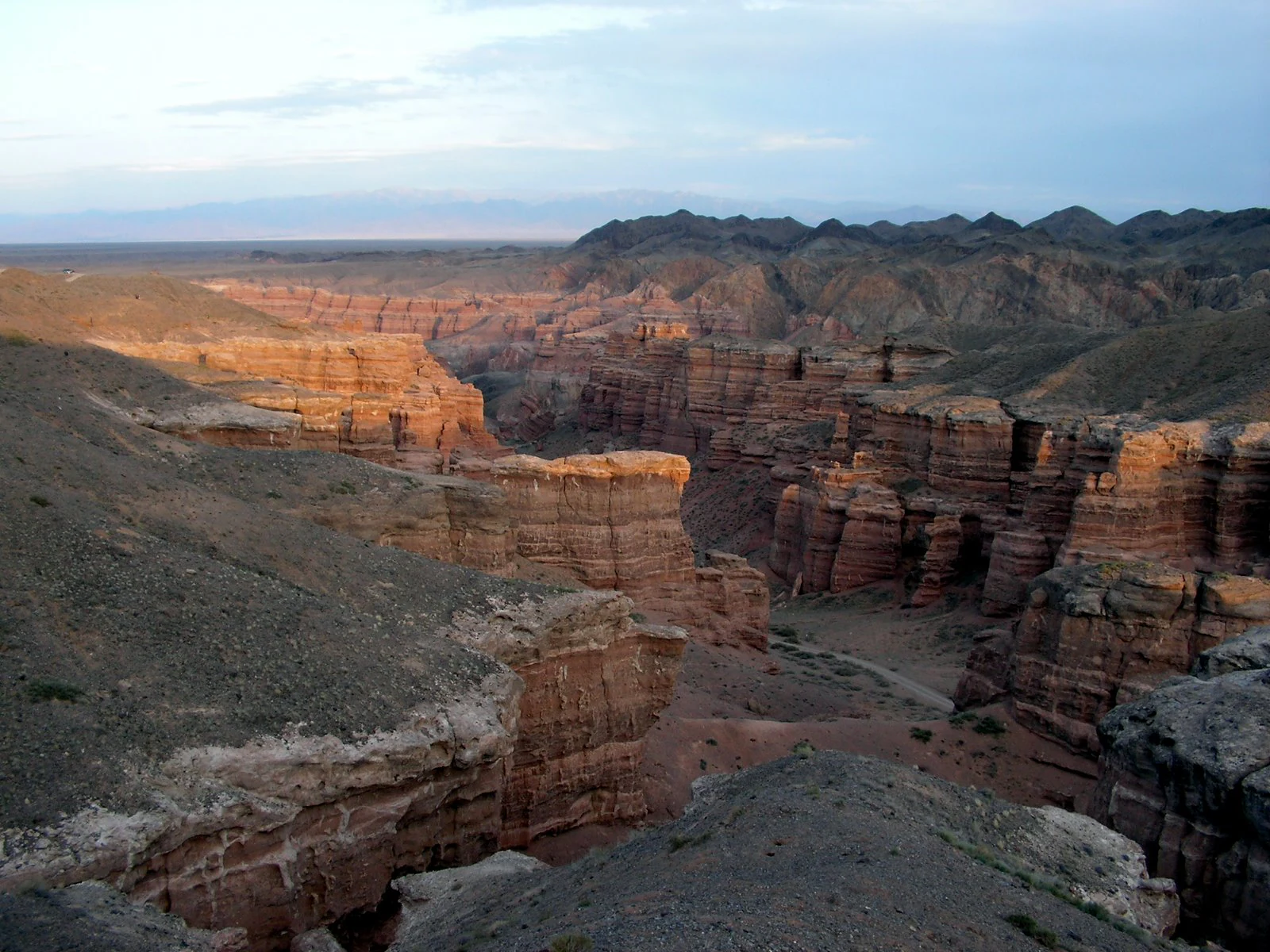 Wide view of Charyn Canyon showing red rock formations, steep cliffs, and distant hills under a partly cloudy sky
