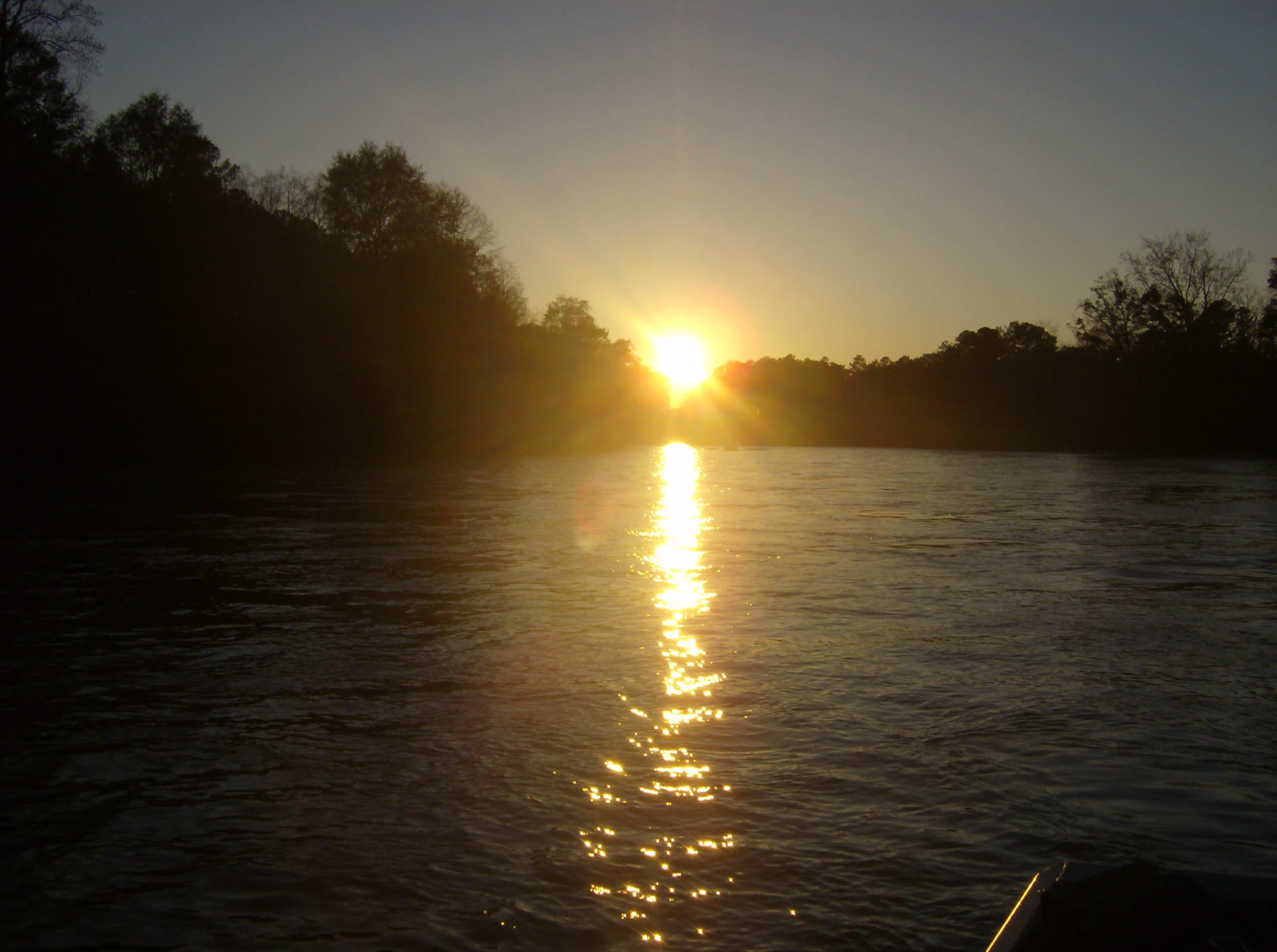 A low-angle, sunlit view across the dark waters of the Chattahoochee River at sunset, with silhouetted trees along the banks.