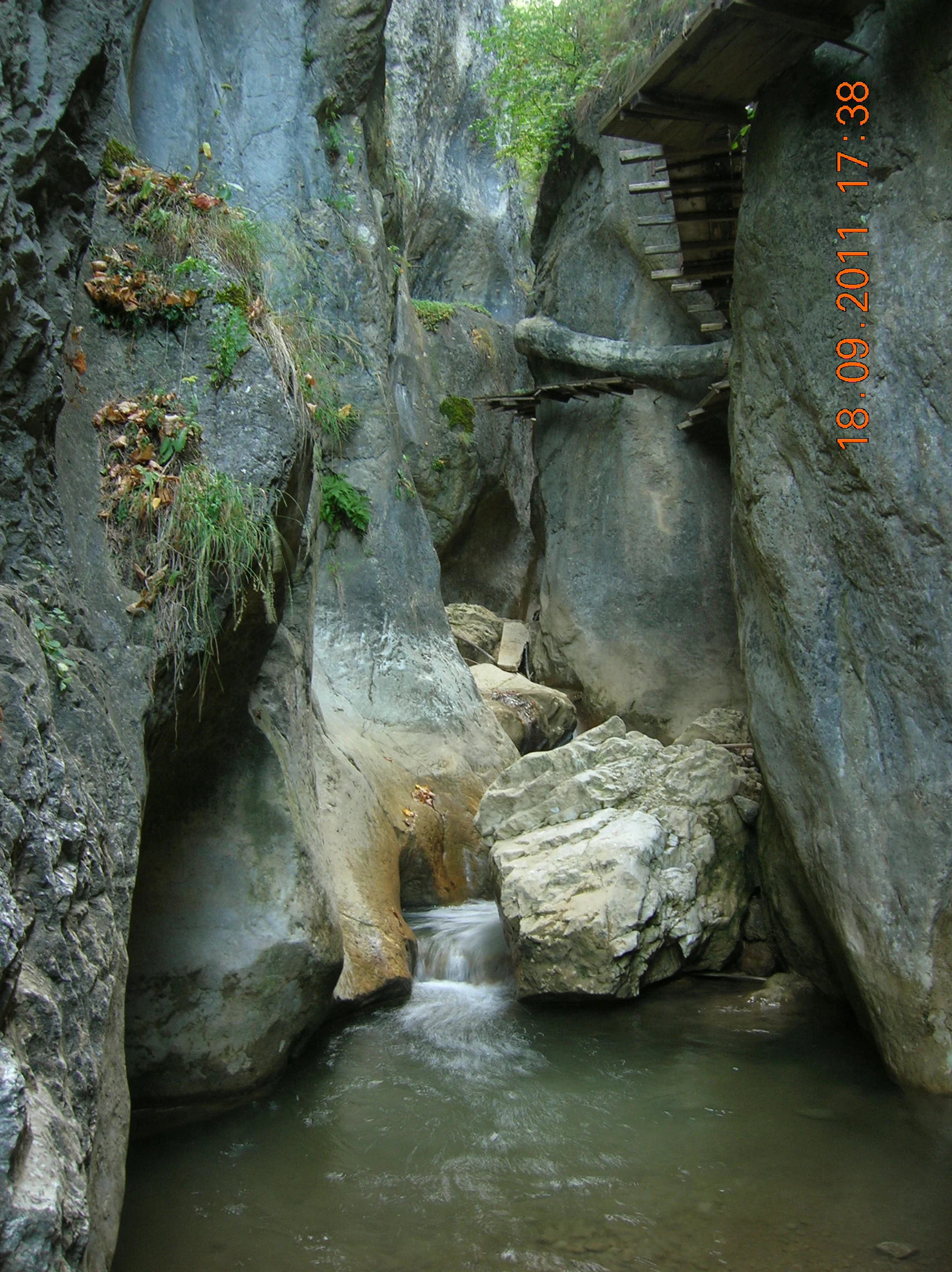 Rocky canyon with a flowing stream, vegetation on rock faces, and a wooden structure on the right side