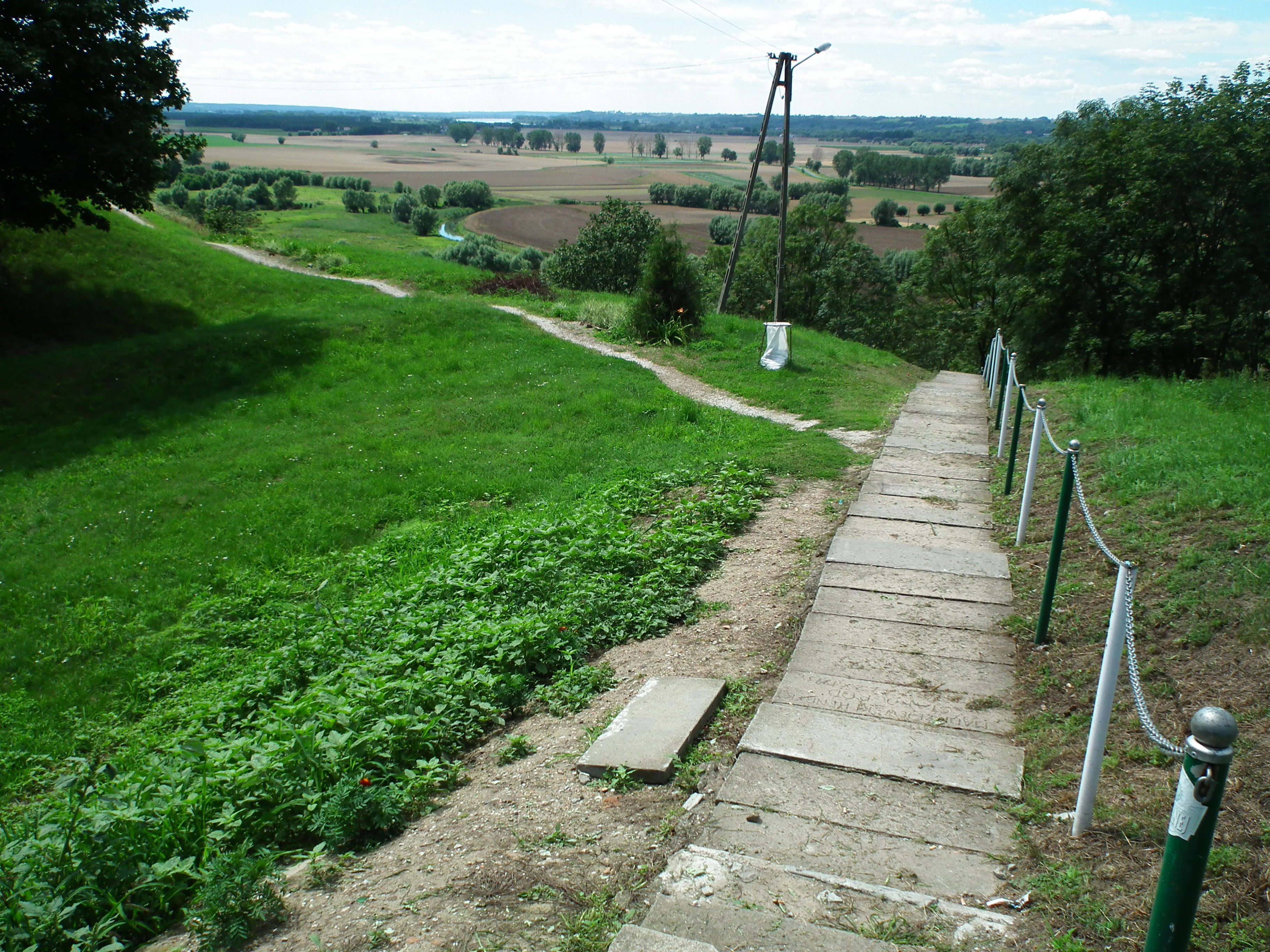 A downward sloping concrete staircase with a rope railing leads from a grassy hill overlooking a valley with fields and trees in Poland.