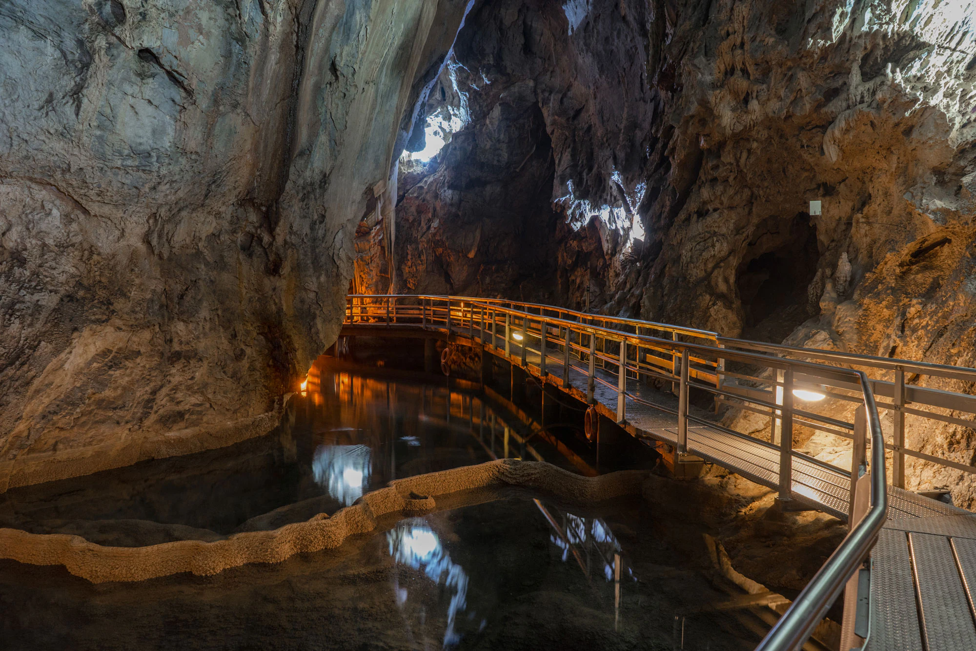 Interior de una cueva de piedra caliza con una pasarela metálica elevada que serpentea sobre una serie de piscinas escalonadas naturales llenas de agua.