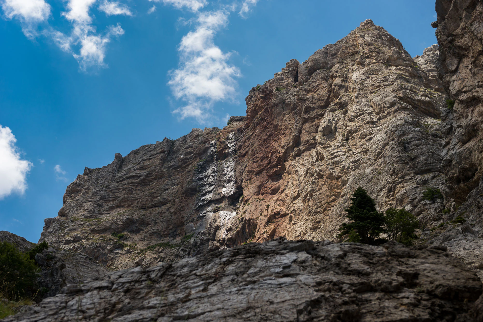 Una cascada estrecha y escalonada que fluye por una cara rocosa empinada y accidentada en las montañas, enmarcada por un cielo azul despejado.