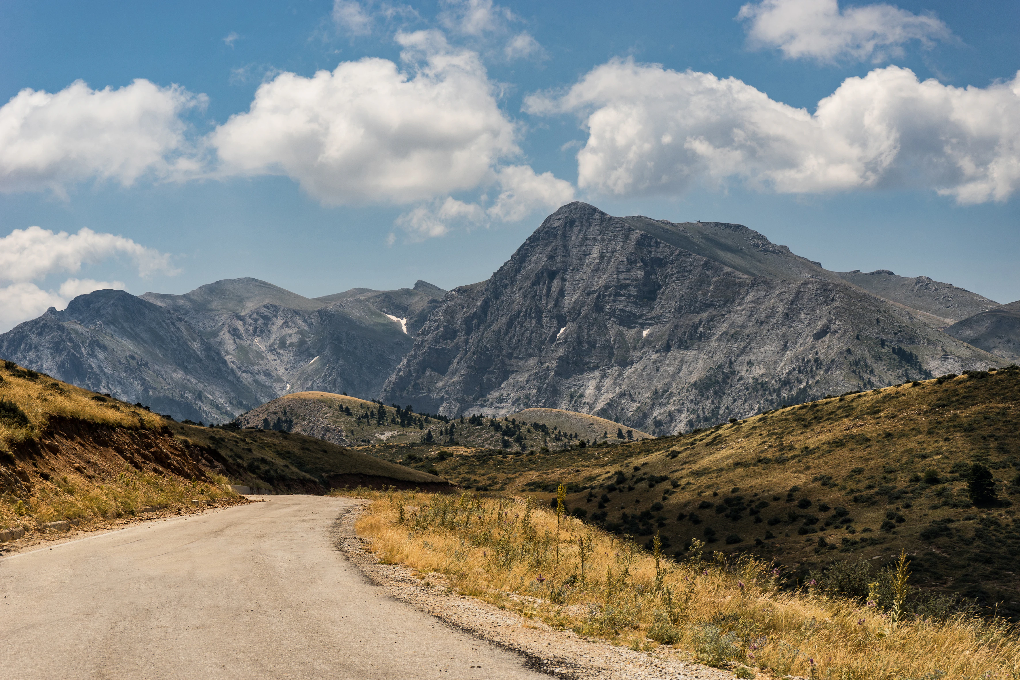 Una carretera sin pavimentar y serpenteante atraviesa un paisaje montañoso seco y cubierto de hierba hacia una cumbre alta y escarpada bajo un cielo azul con nubes dispersas.