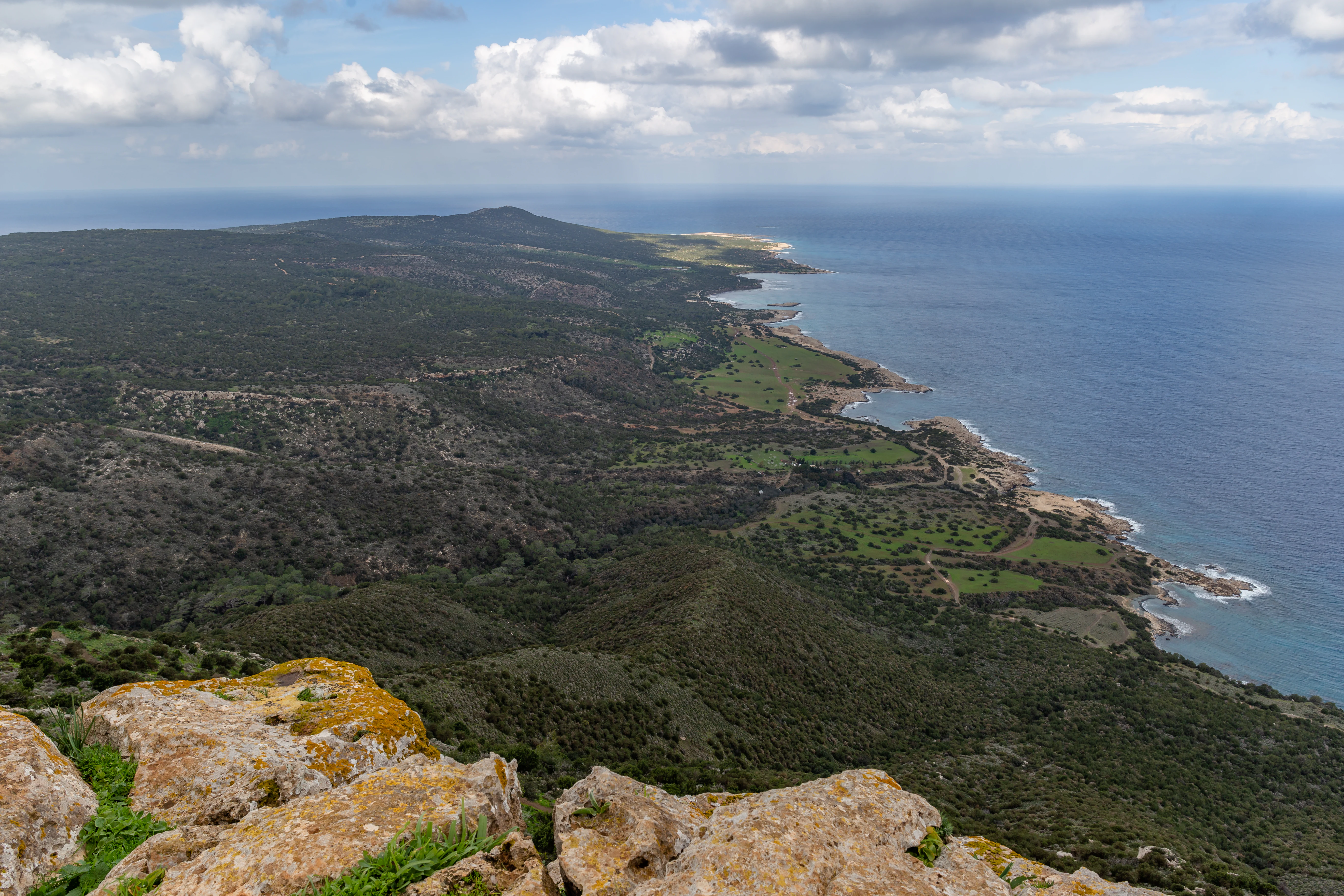 A high vantage point view looking down from a rocky summit over the rolling green hills and coastline of the Akamas Peninsula in Cyprus, surrounded by the blue Mediterranean Sea under a cloudy sky.