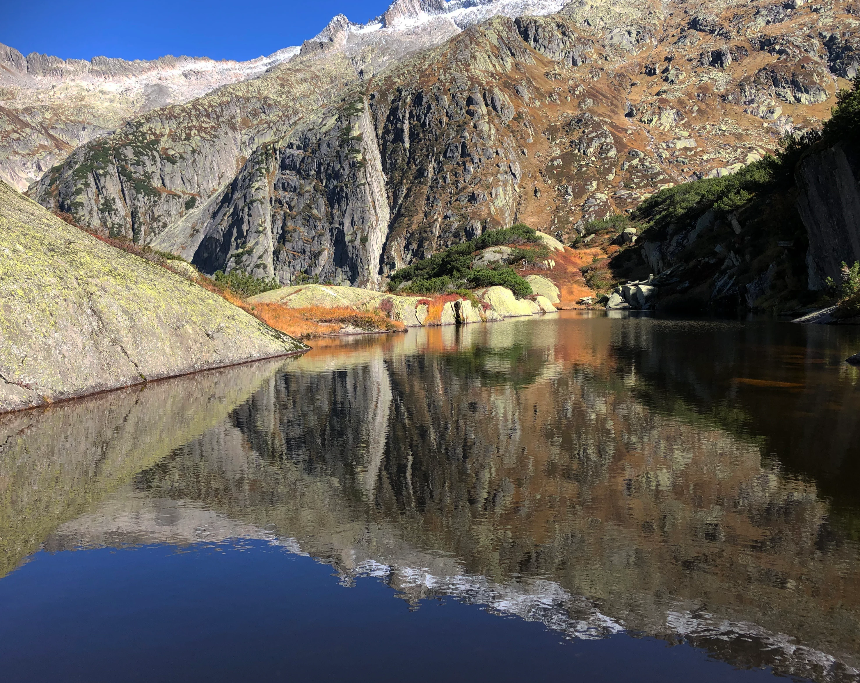 A small, calm pool of water reflecting the surrounding rugged mountain slopes and clear blue sky.