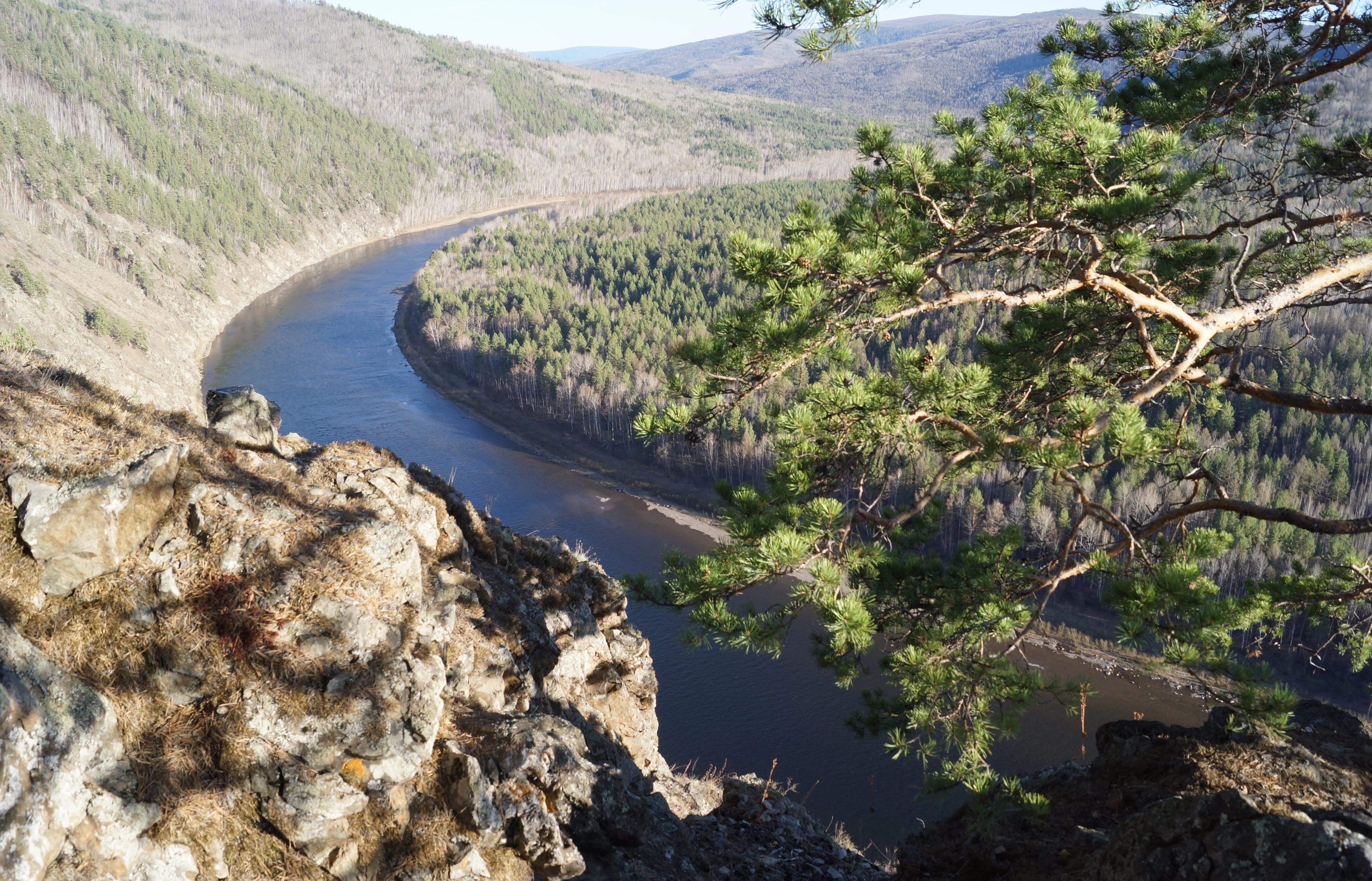 A wide river flowing through a forested valley with rocky terrain in the foreground and rolling hills in the background