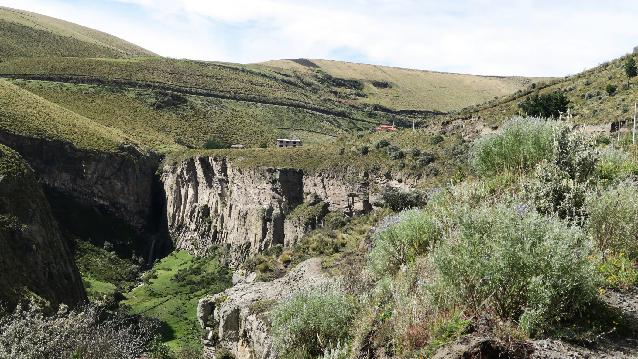 A deep rocky canyon with steep walls cuts through rolling, grass covered hills, with small structures visible on the distant horizon under a cloudy sky.