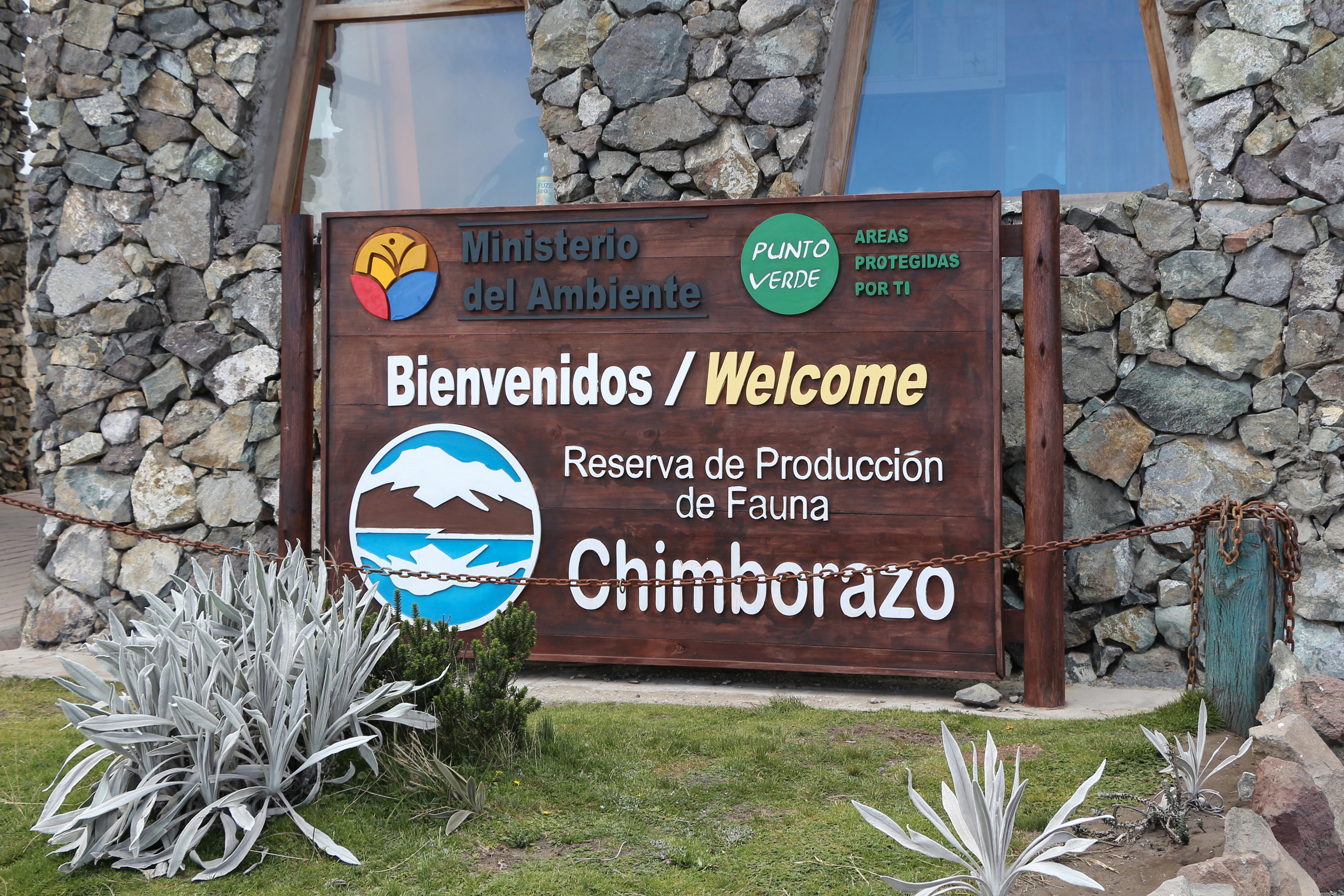 A wooden sign with text in Spanish and English reading Bienvenidos / Welcome, Reserva de Produccion de Fauna Chimborazo, mounted on a stone wall exterior.