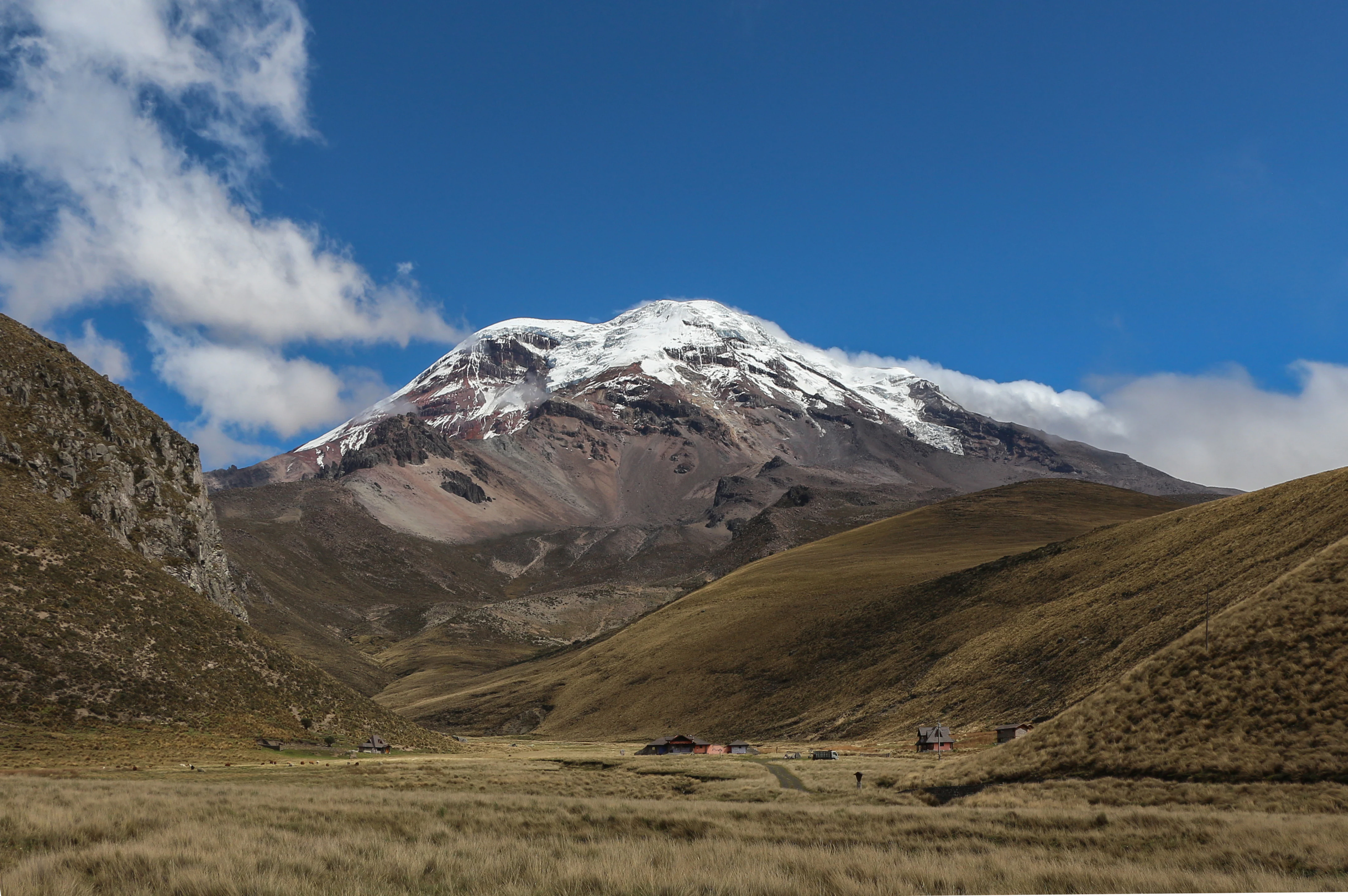 A wide view of the snow-covered, multi-peaked Chimborazo volcano towering over a vast, dry grass-covered valley in the Andes.