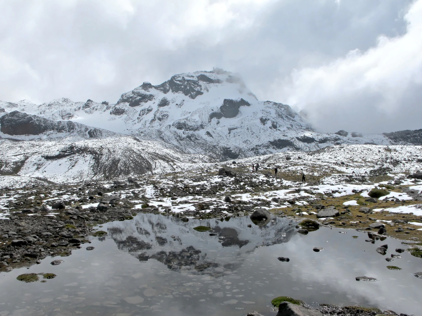 A snowy mountain peak with a rocky, high-altitude landscape in the foreground, including a small pond that reflects the mountain.