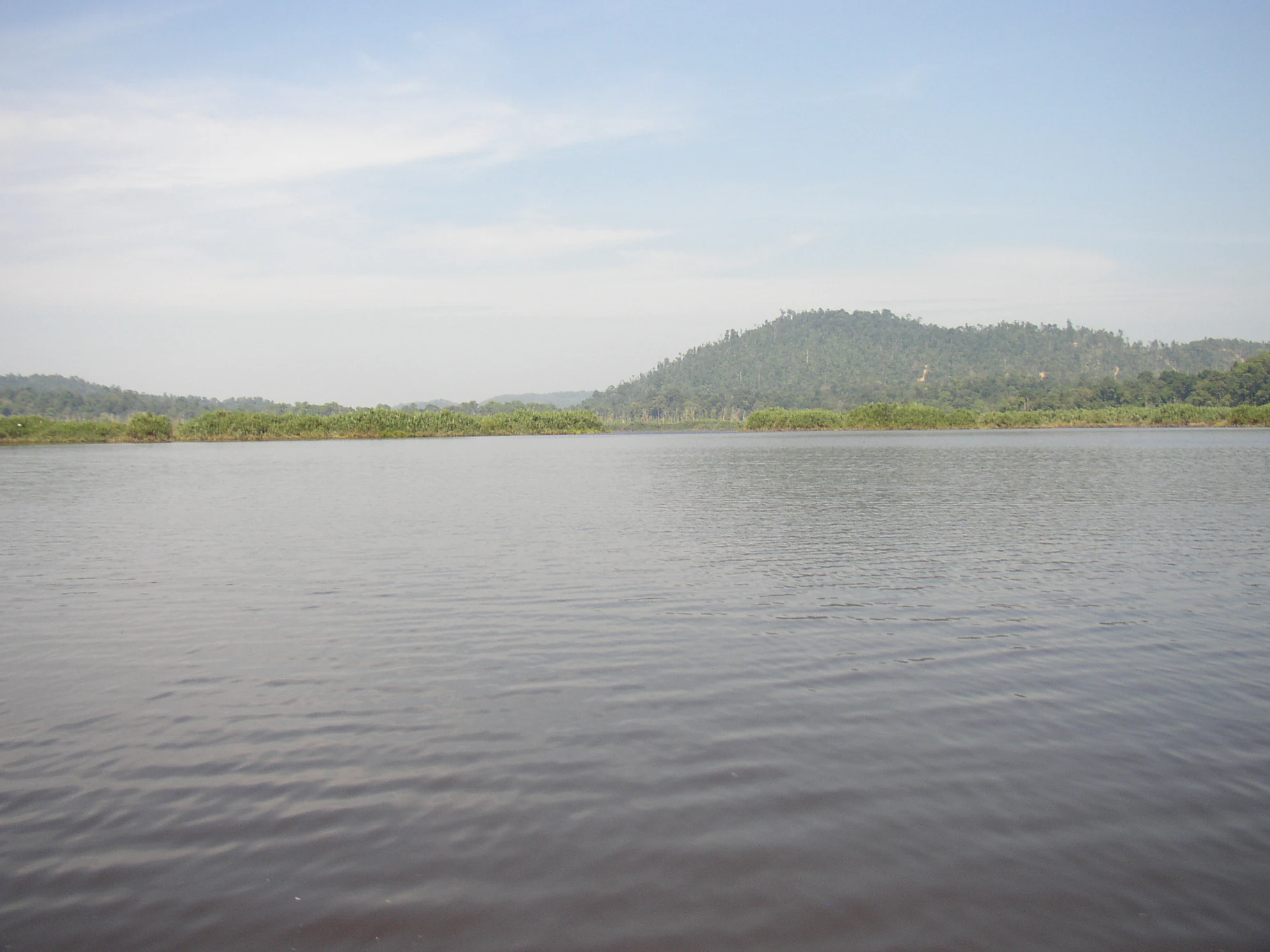 A wide view of Chini Lake showing dark, calm water in the foreground and tree-covered hills in the background under a hazy sky.