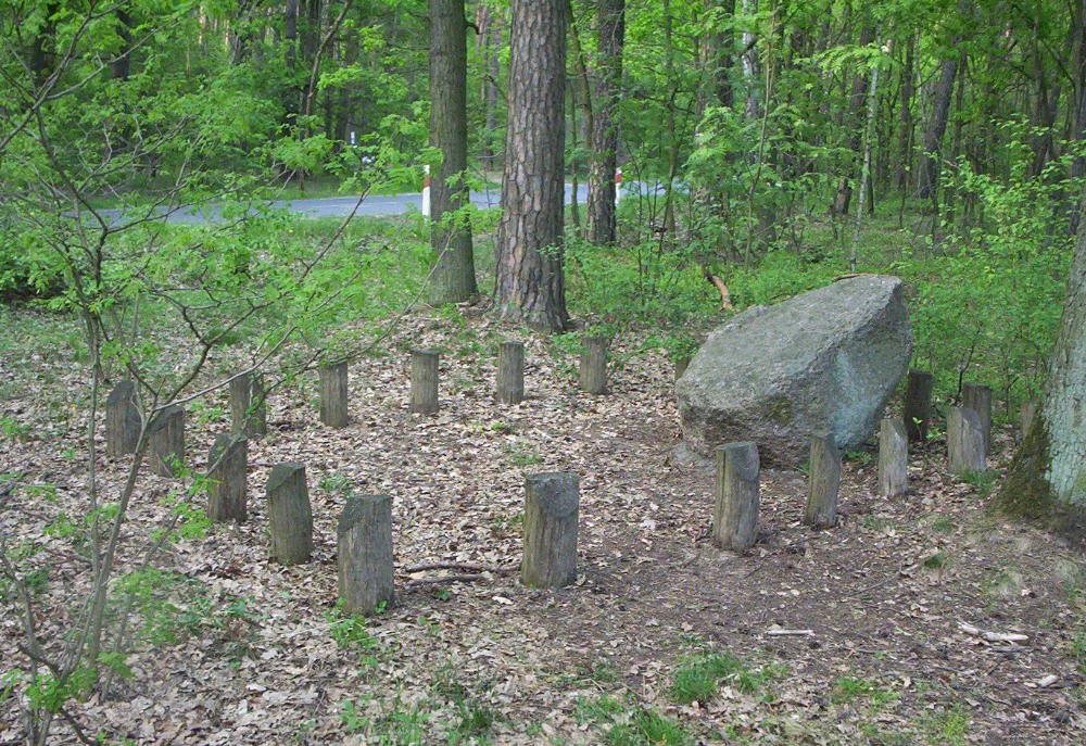 A large grey boulder sits on a forest floor, partially encircled by a ring of short wooden posts in a wooded area near a road.