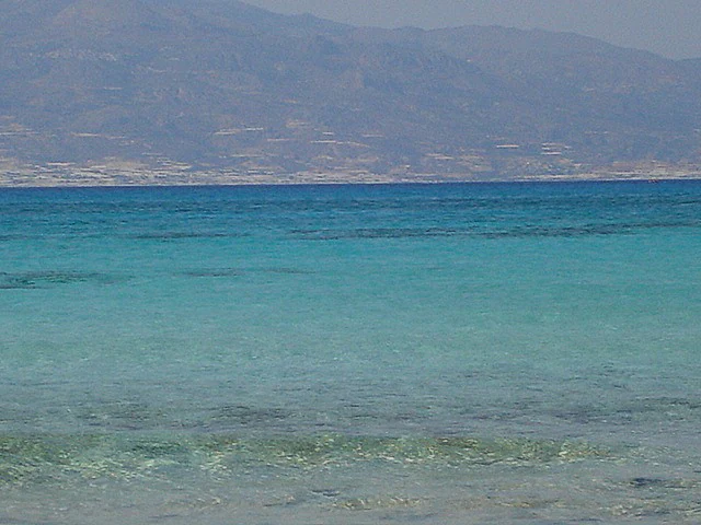 A horizontal view of bright turquoise sea water in the foreground, with a distant, hazy island coastline and mountain range in the background under a pale sky.