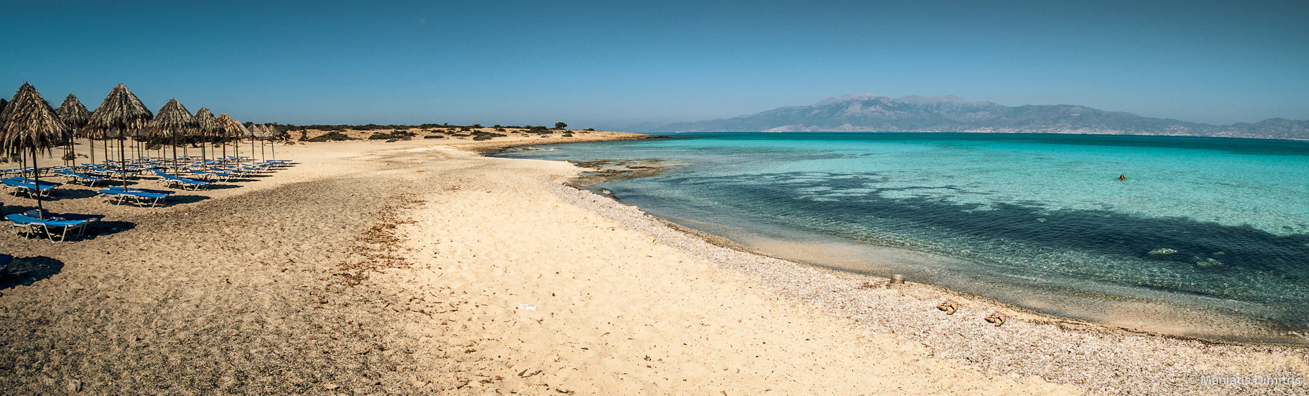 A wide panoramic view of a sandy beach on Chrysi Island with rows of thatched sun umbrellas and lounge chairs on the left, leading to shallow, clear turquoise water, with a faint coastline visible in the background under a clear sky.
