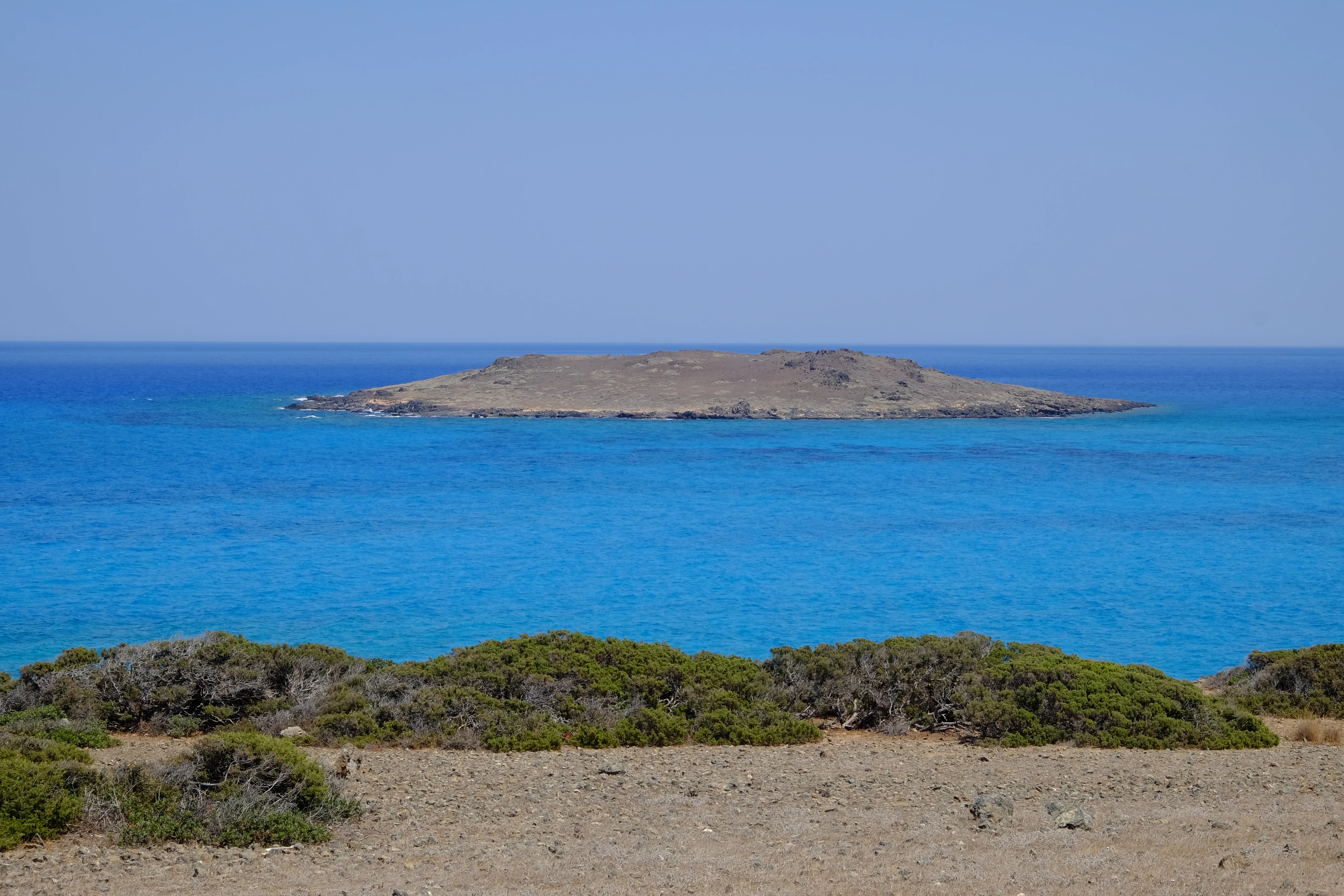 A distant small, flat island surrounded by vibrant turquoise water, seen from the rocky, arid terrain of a foreground shoreline.