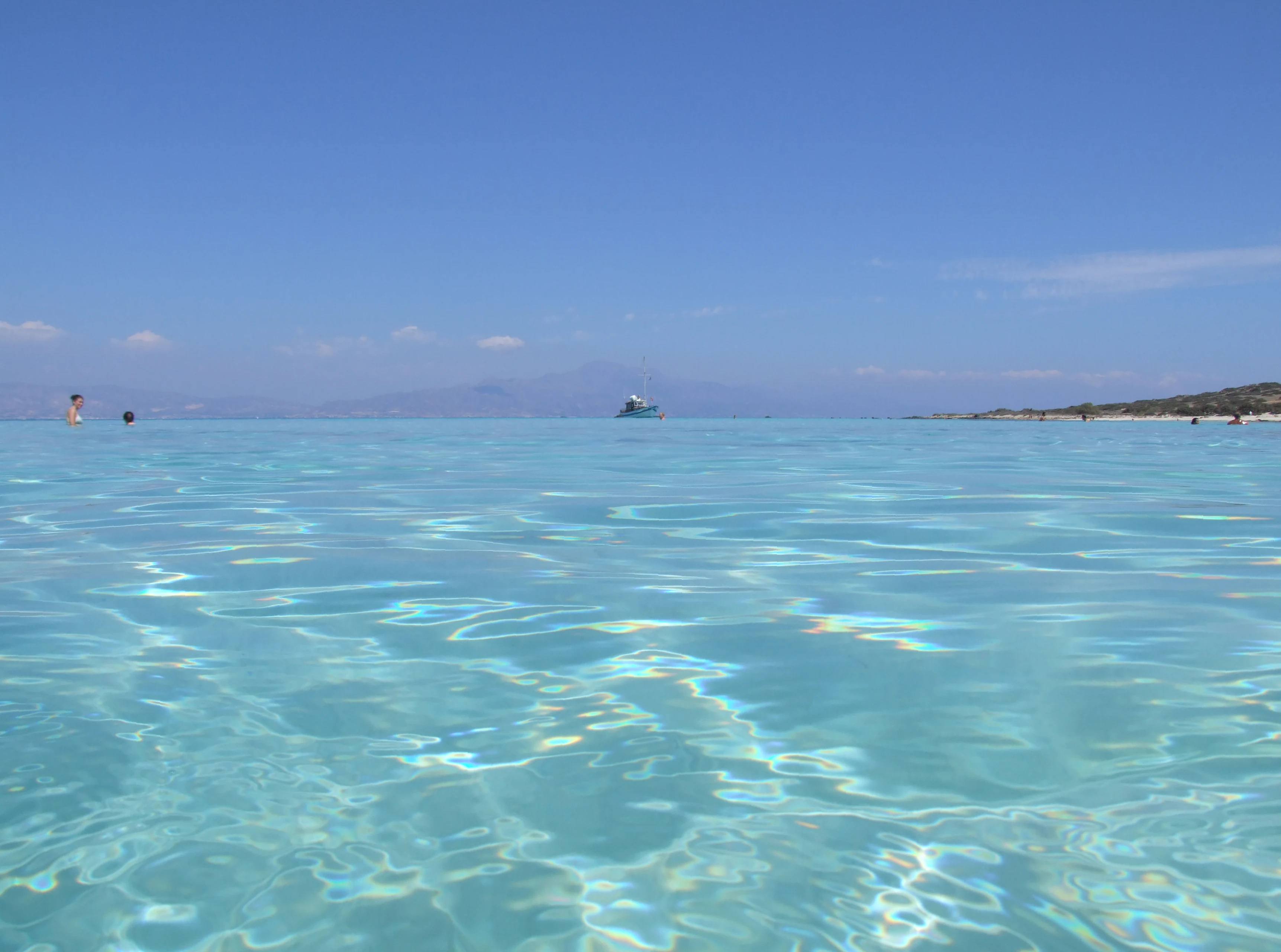 Eye-level shot of clear, shallow, light blue ocean water at Chrysi island under a bright blue sky, with a distant coastline visible.