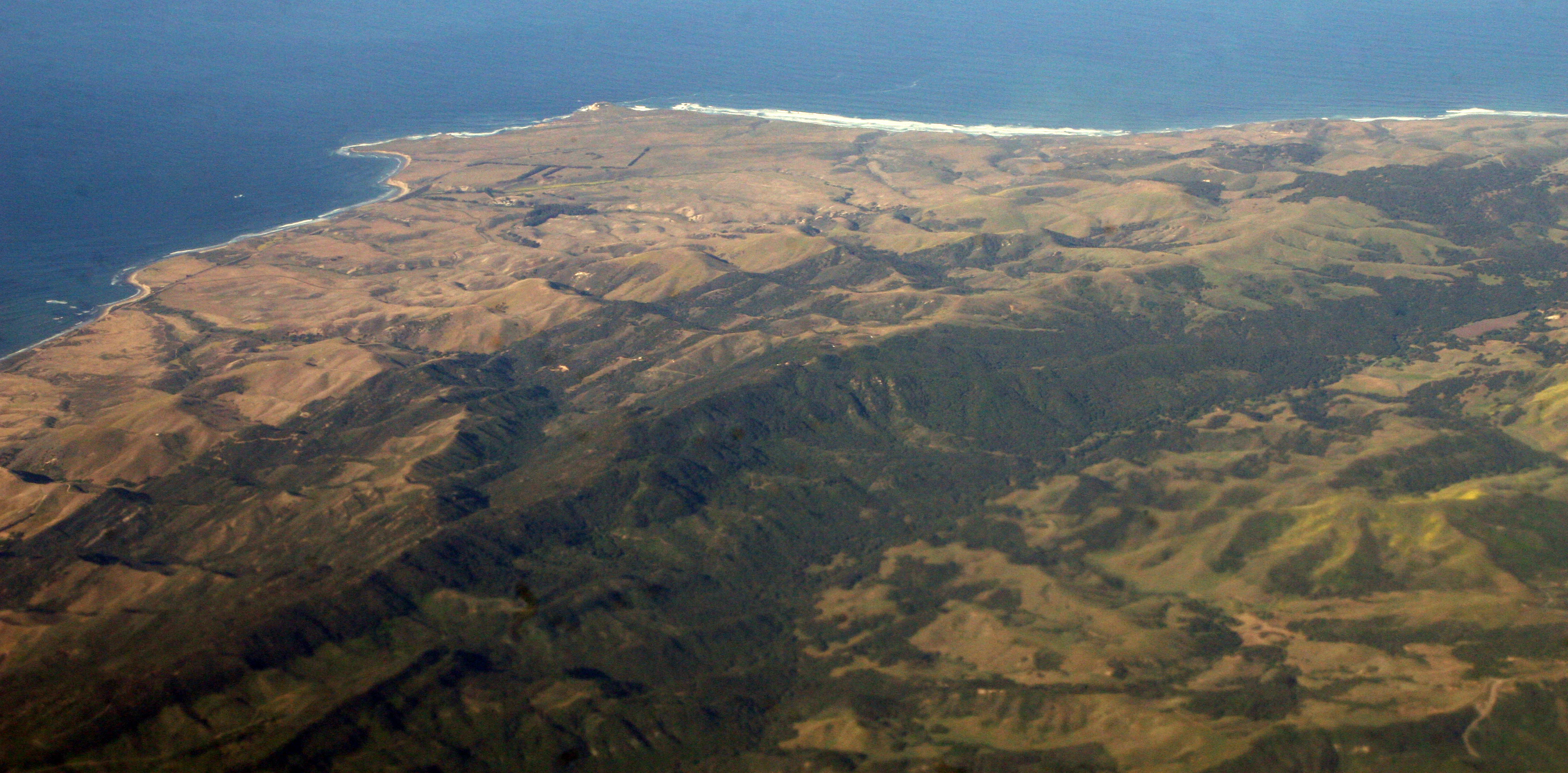 An aerial view of the California coastline showing the prominent point of Point Conception curving into the Pacific Ocean, backed by the rolling terrain of the Santa Ynez Mountains.