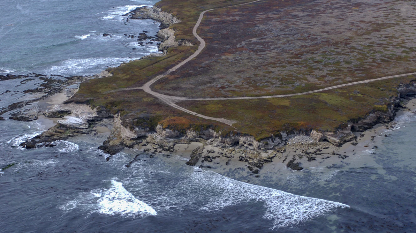 Aerial view looking down at a rocky coastline with a dirt road winding across the headland into the Pacific Ocean.
