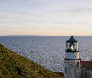 A white lighthouse standing on a grassy coastal bluff next to the open Pacific Ocean at twilight.