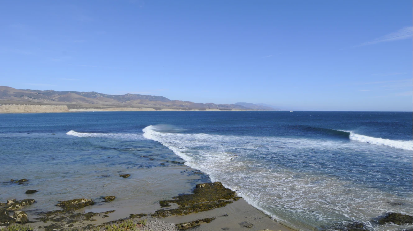 A wide view of the California coast featuring breaking ocean waves in the foreground, a rocky shoreline, and a distant headland under a clear blue sky.