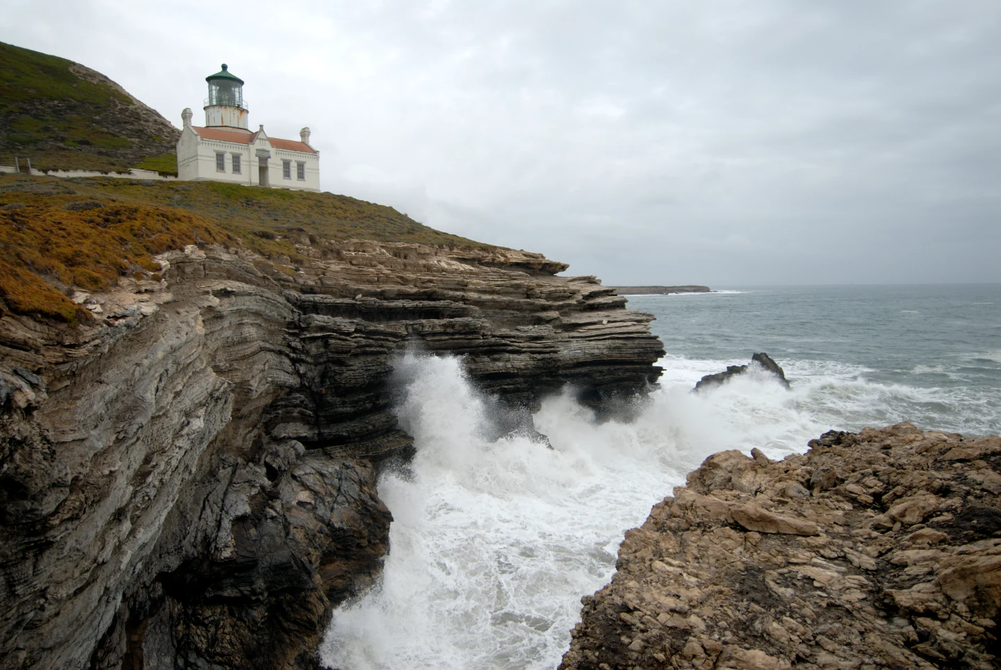 A white lighthouse stands on a rugged cliff overlooking a rocky coastline where ocean waves crash against the shore under an overcast sky.