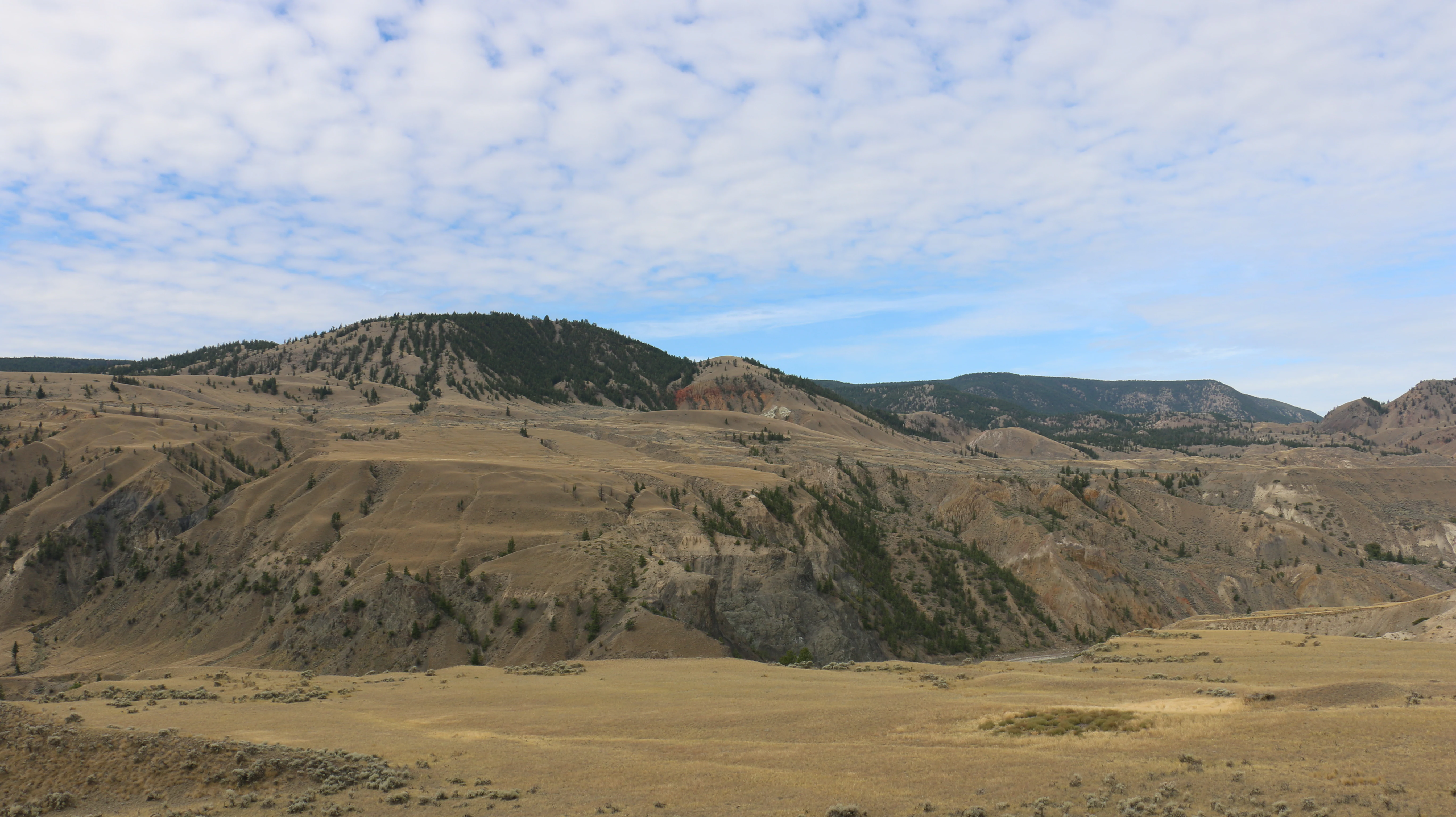 Wide landscape view of dry rolling hills and canyon terrain with sparse vegetation, rocky outcrops, and a partly cloudy sky.