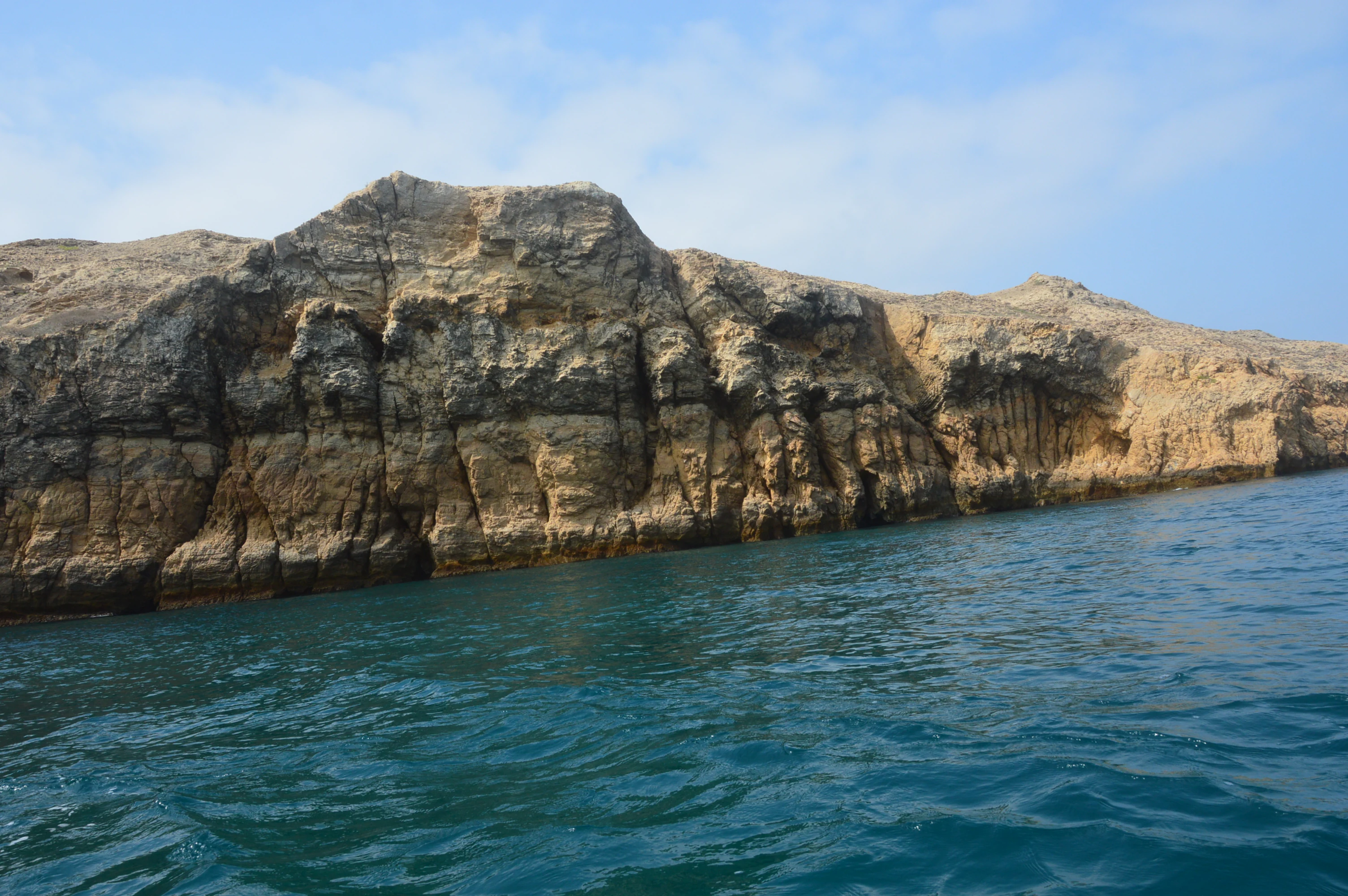 A low-angle view of the steep, light-brown, stratified cliffs of Churna Island against a bright sky, meeting the turquoise water of the Arabian Sea.
