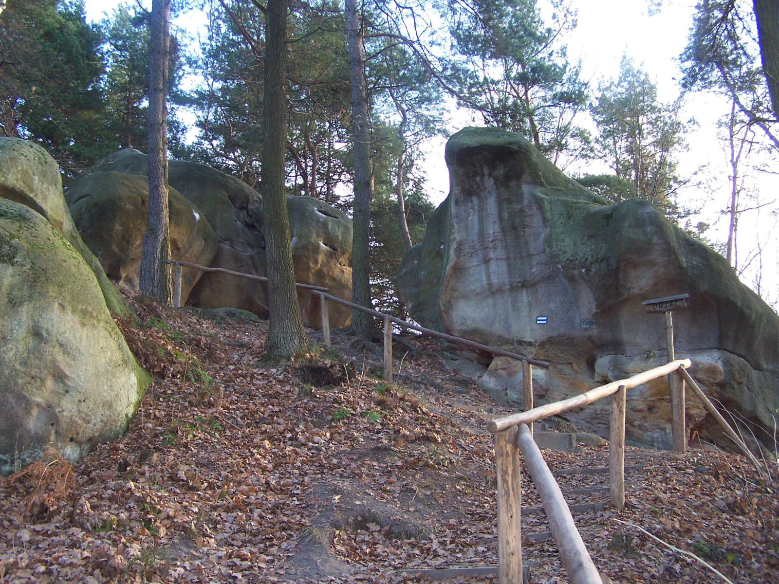 A dirt path winds between large, weathered sandstone rock formations in a forest, with a simple wooden railing protecting the trail.