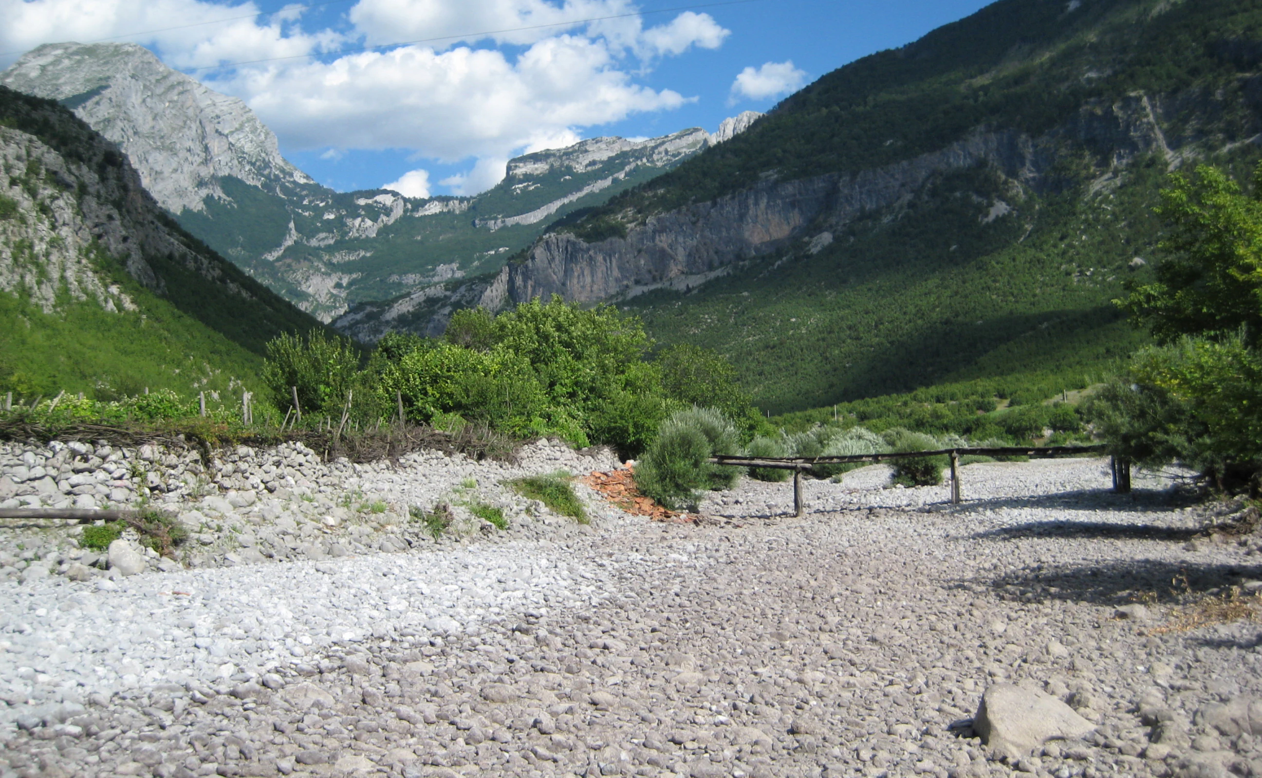 A wide, dry, rocky riverbed stretches toward a lush green mountain valley under a blue sky with scattered clouds.