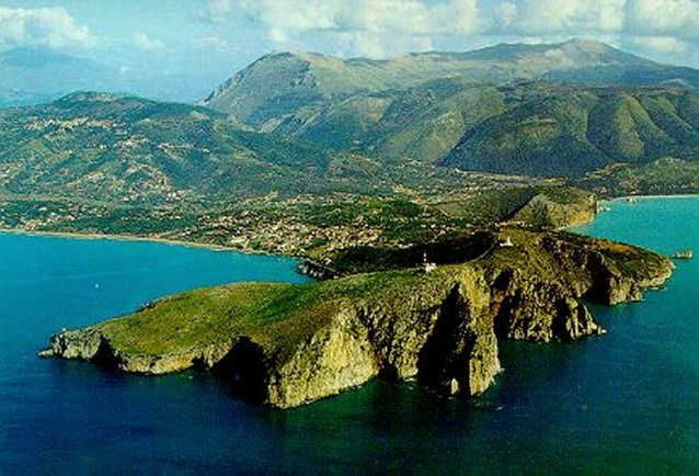 Aerial view of a coastal headland with blue water on both sides, green hills, and mountains in the background.