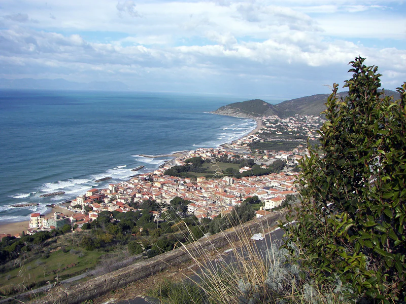 Panoramic view of Santa Maria di Castellabate town with red-roofed buildings along the coastline, ocean waves, and surrounding hills under a partly cloudy sky